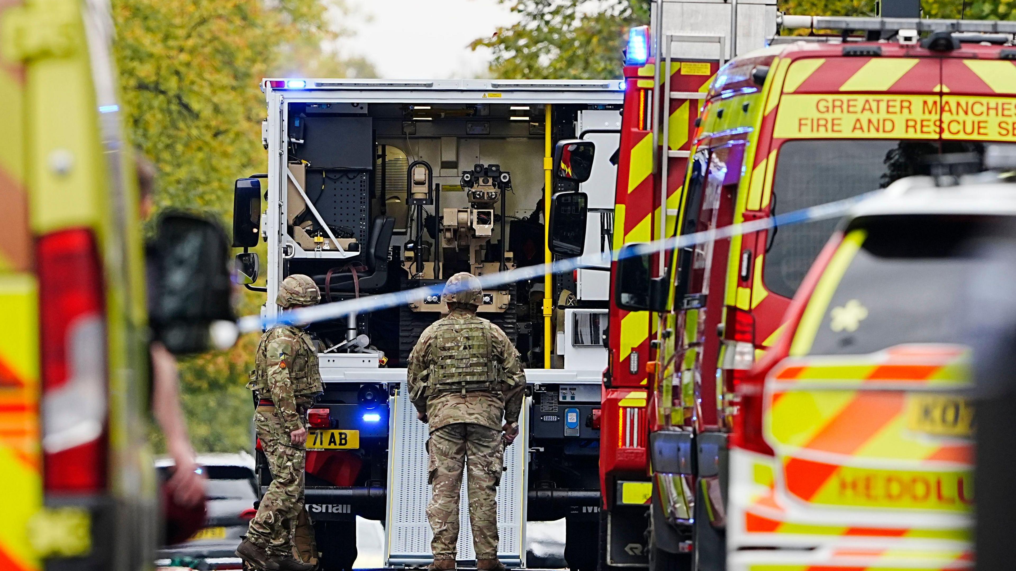 Members of the army in military fatigues stand outside a van with a bomb disposal robot seen inside. Police vehicles and police tape are seen in the foreground. 