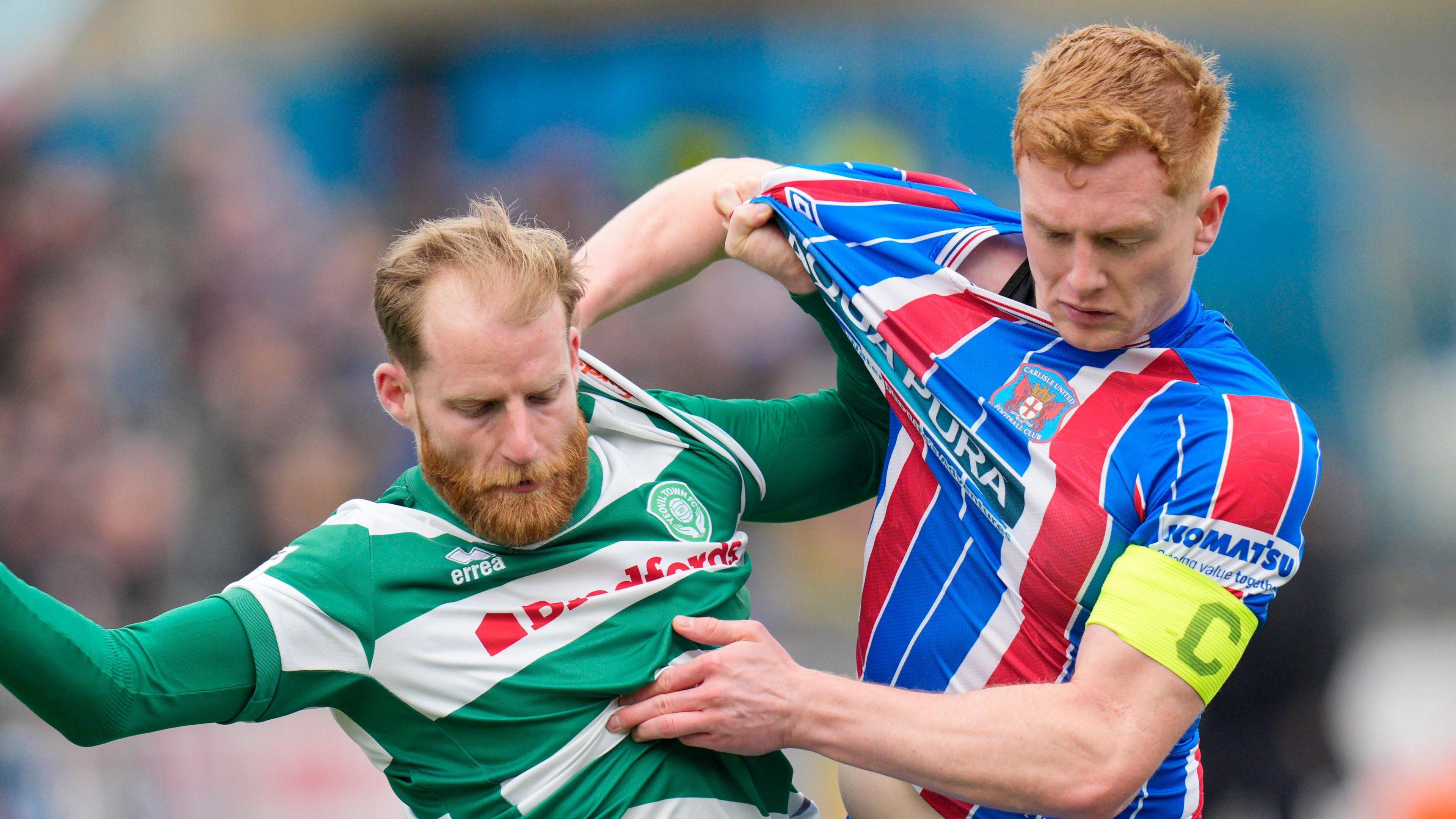 Morgan Feeney (right, wearing a captain's armband with 'C' on it) is grabbling with Yeovil player Josh Sims as they pull each other's shirts and have their eyes looking down at the ball