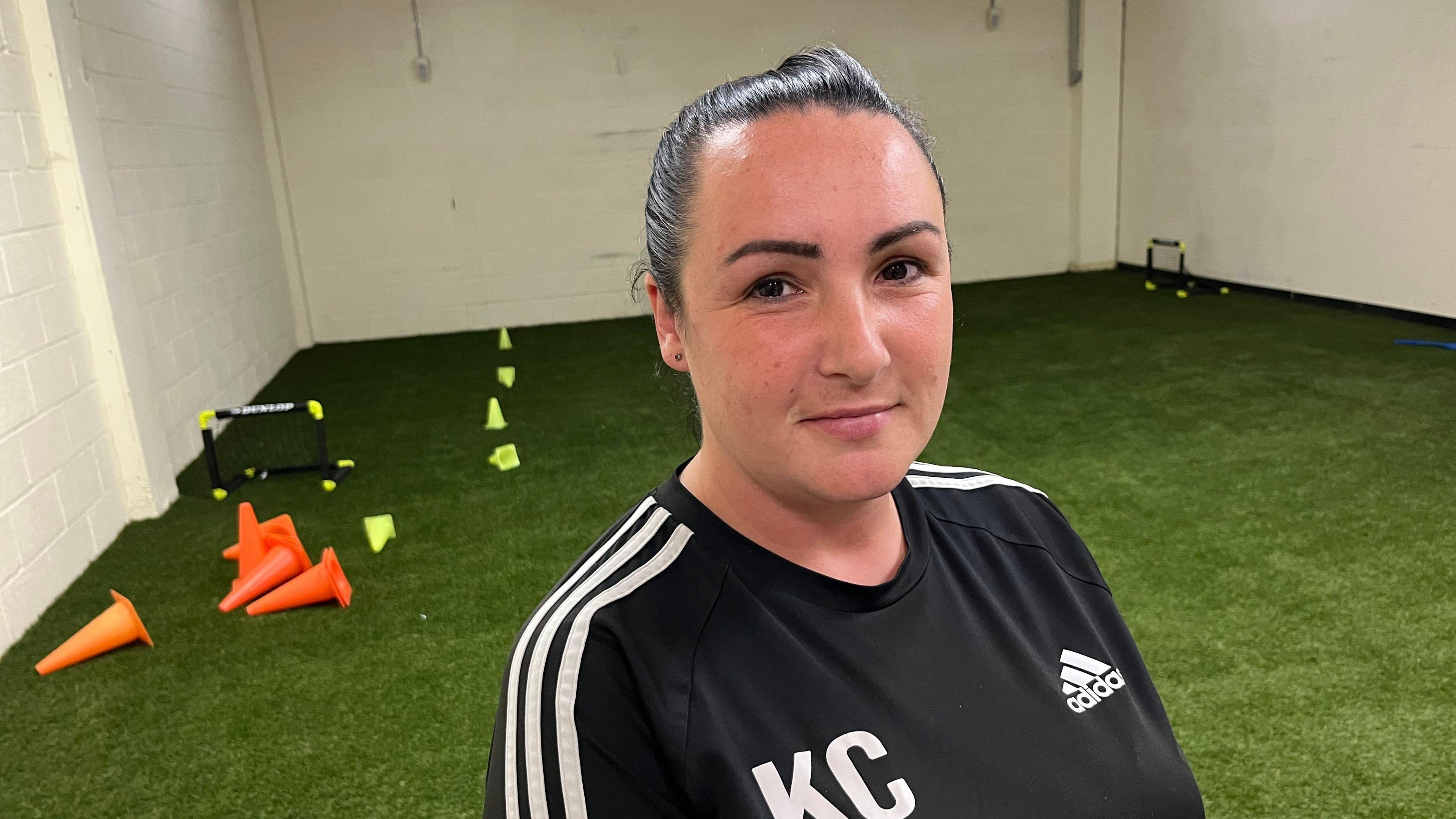 Kerry Cassidy is looking at the camera. She is wearing a black top with an Adidas logo and stripes and the initials KC on the front. Behind her is an indoor pitch with artificial turf, cones and small nets.