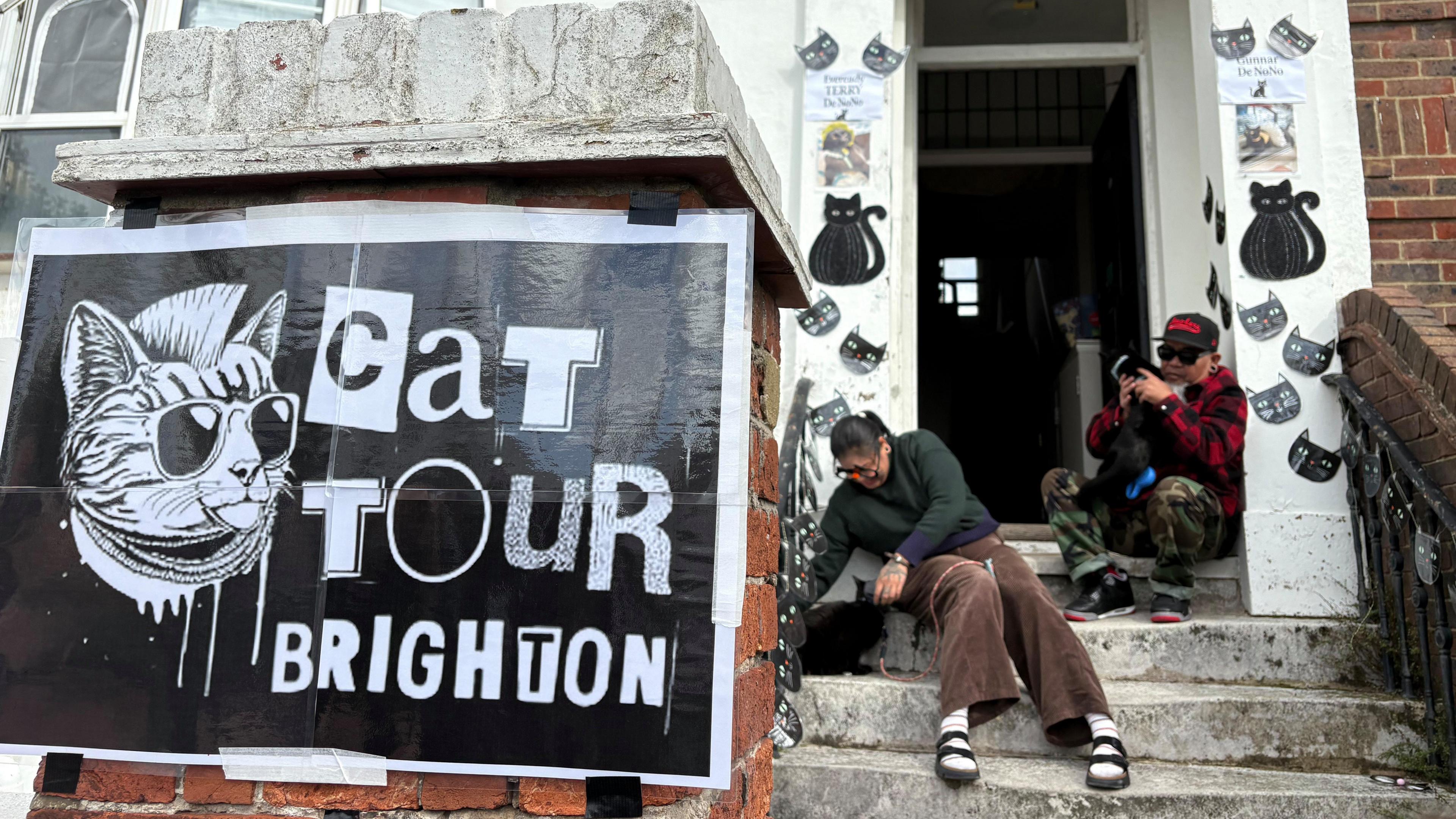 Two cat owners and volunteers for Brighton Cat Tour sit on concrete steps outside their flat. On the red brick column in front of the steps and doorway sits a punk-themed poster reading "Cat Tour Brighton", depicting a cat with a mohawk, sunglasses and chains sticking its tongue out. The owners sit and pet two black cats, with black cat-themed decorations adorning the front entryway to their home for passing visitors.
