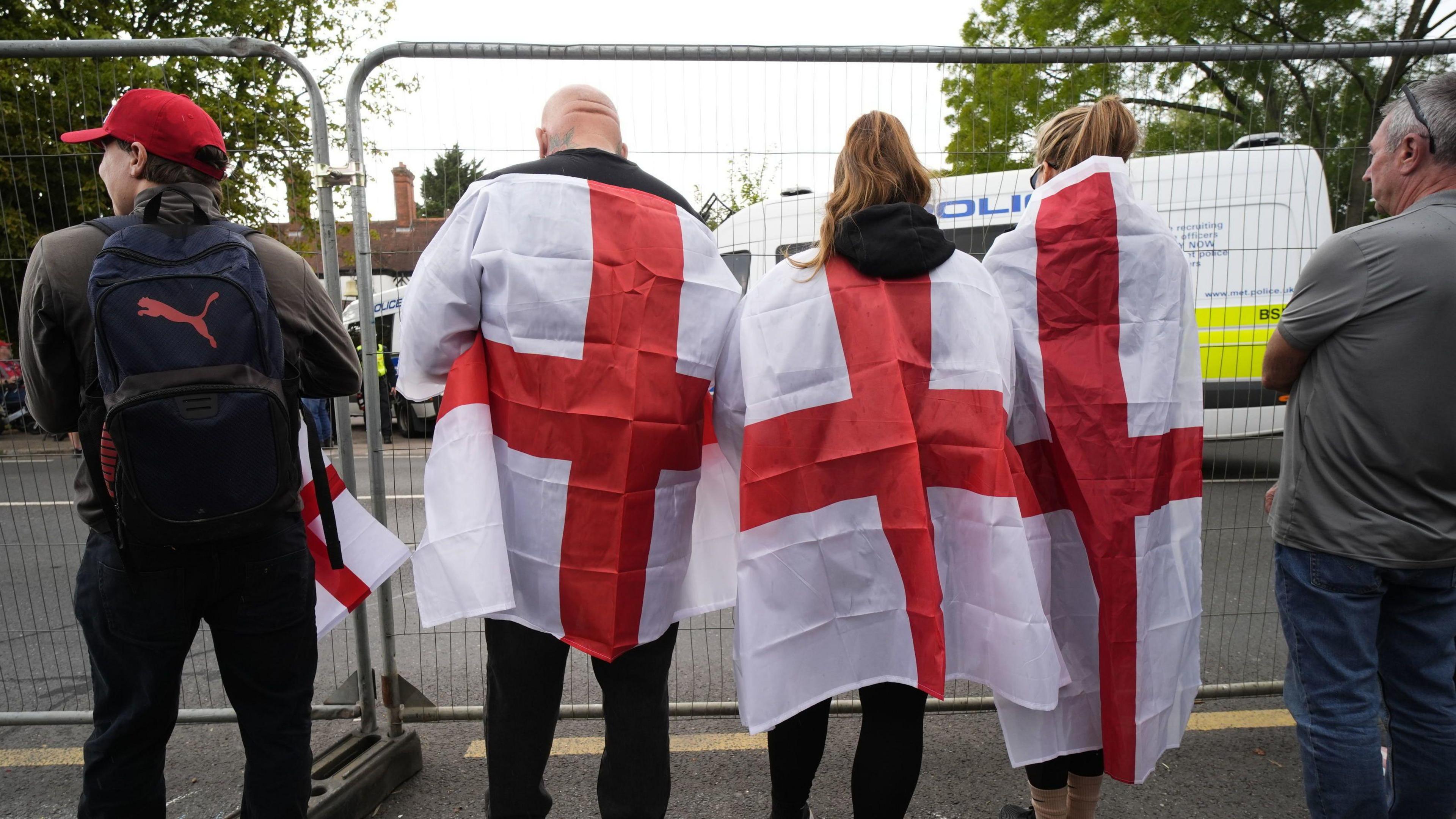 Five people, three of whom have the flag of St George draped round their shoulders, standing behind a metal fence. In front of them is The Bell Hotel, which is blocked off by police vans.