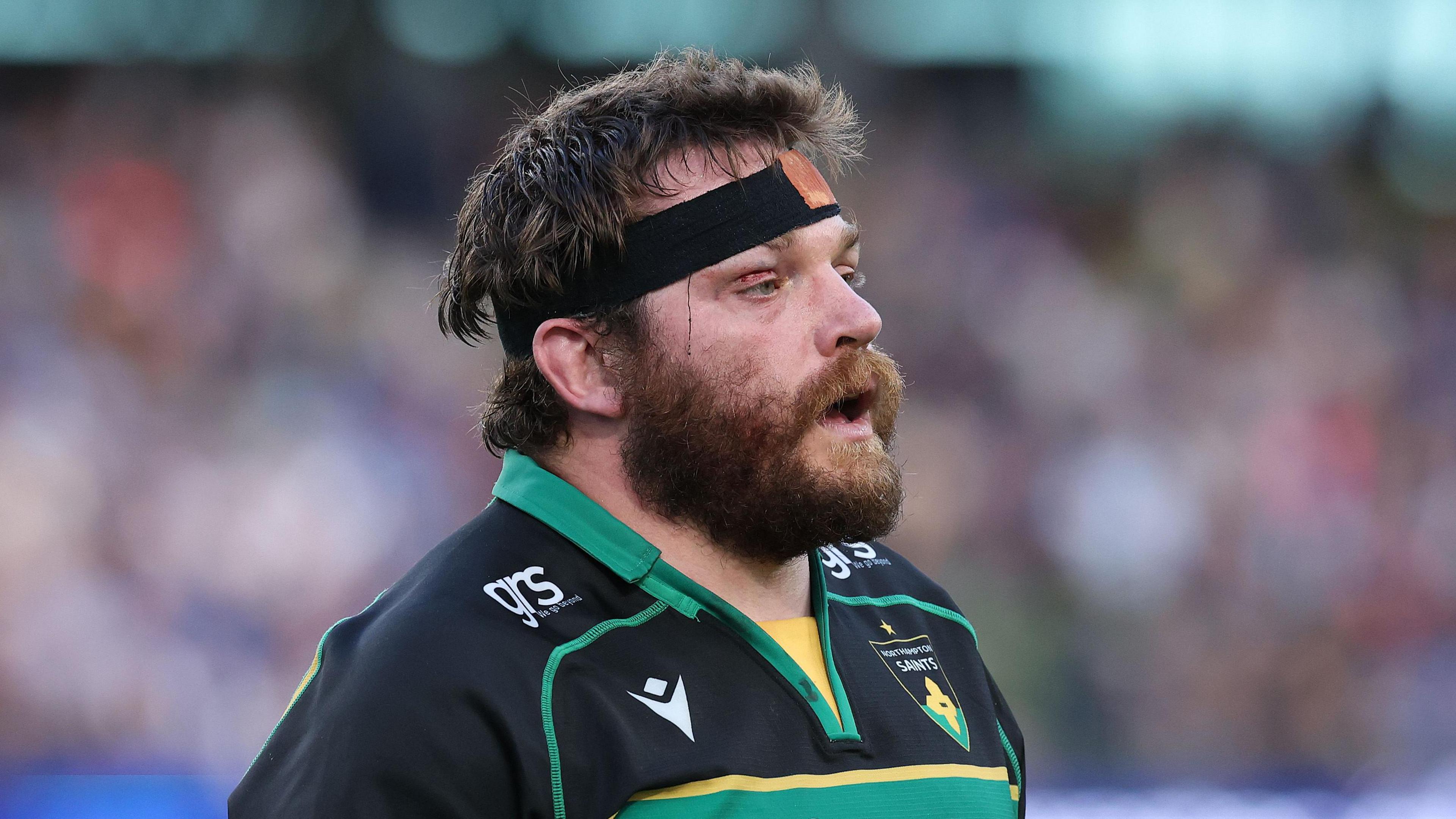 Elliot Millar Mills playing for Northampton Saints. He is wearing a headband and has a thick browny-gingery beard and has a slight cut on his right eye