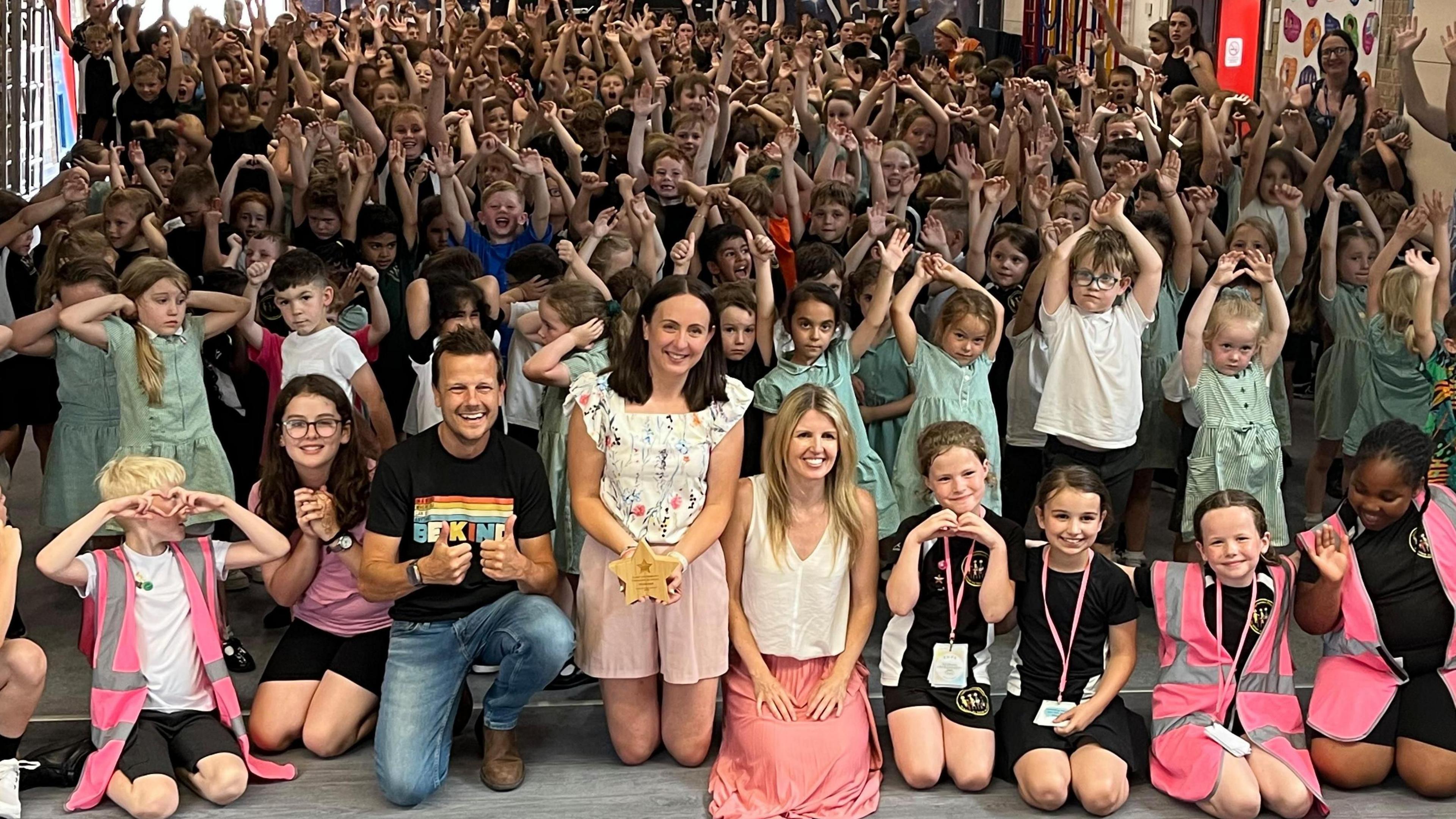 A hall full of children with their hands in the air cheering with three adults at the centre front