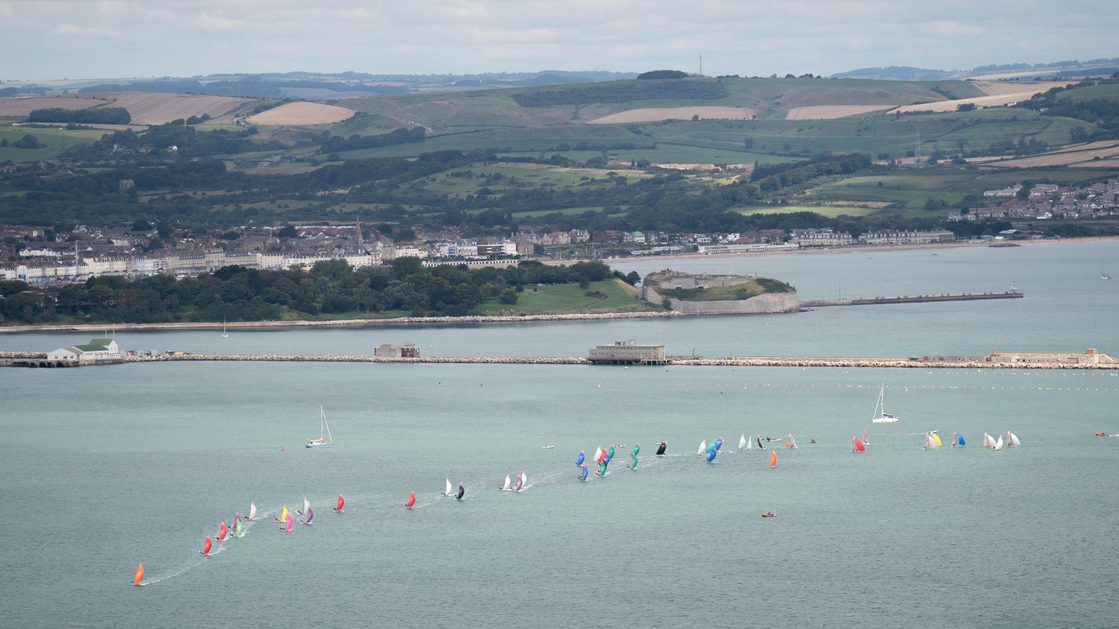 View of Portland Harbour with Weymouth in the background. A long line of small boats with different coloured sails runs from right to left across the picture.