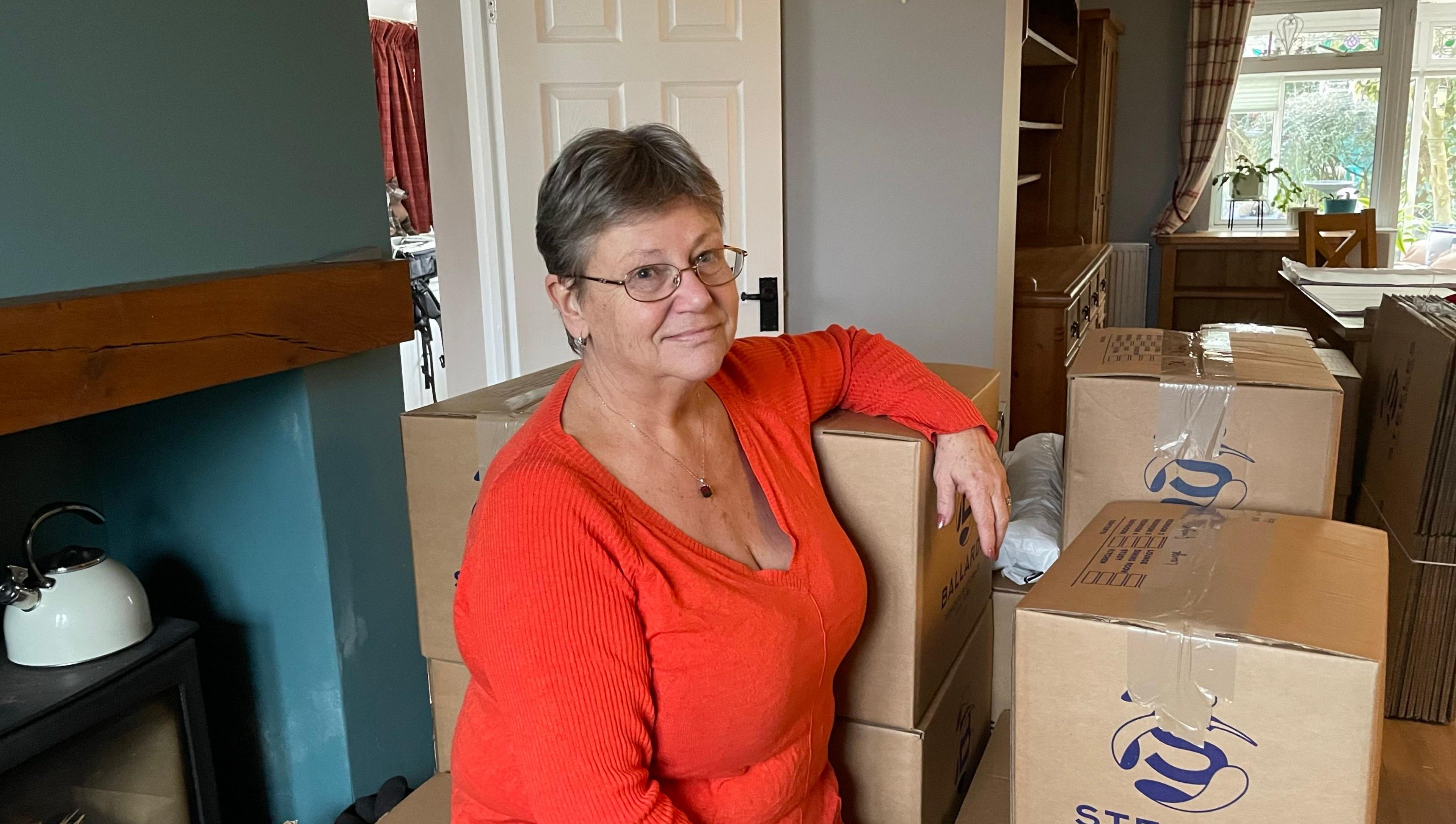 Jayne Keeley is wearing a red/orange long sleeved top, has short brown hair and is wearing glasses. She is standing in a room surrounded by cardboard boxes containing possessions that are going into storage. A fireplace with a teal coloured chimney breast is on the left of the image.