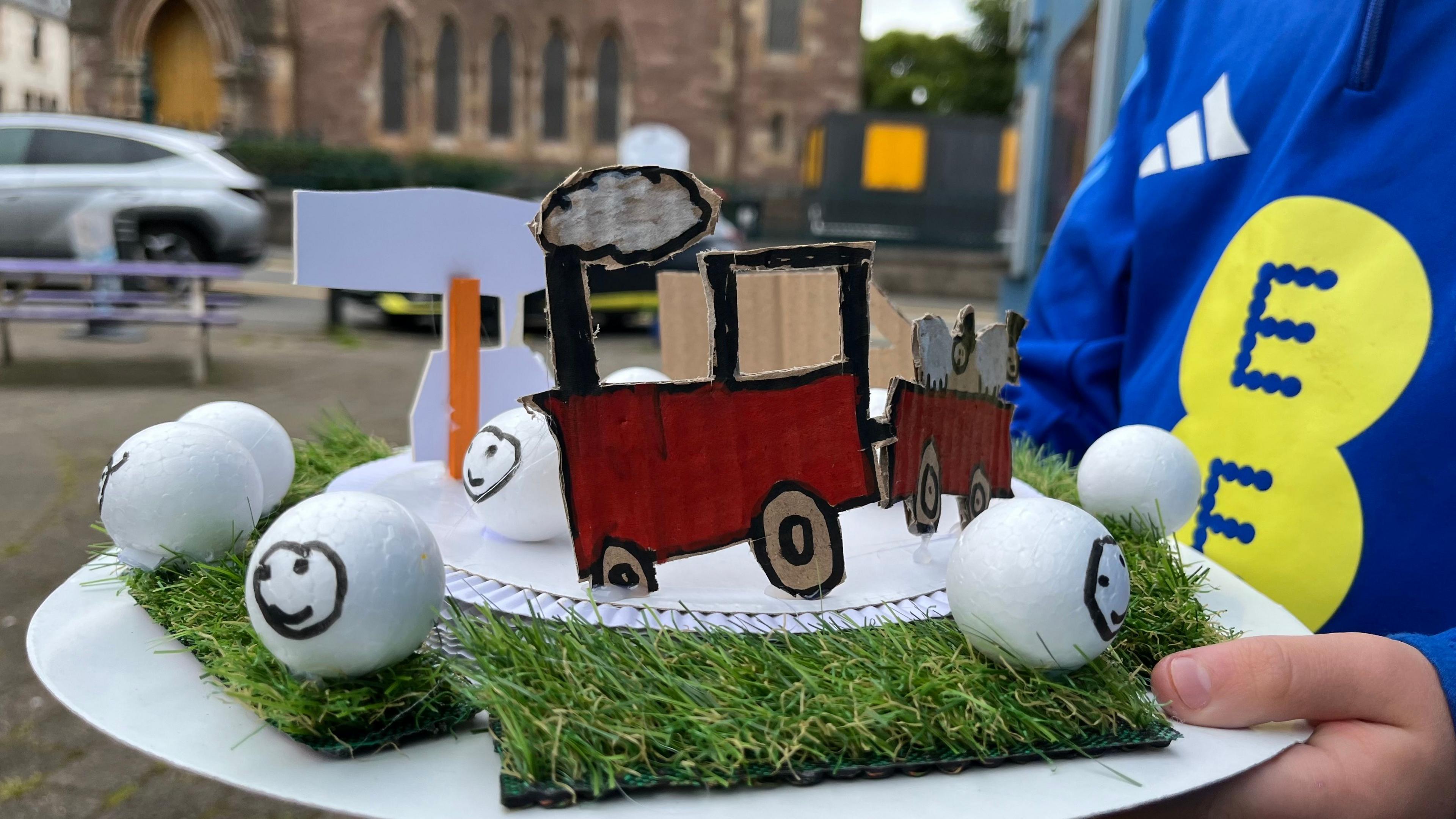 Dealbh a' sealltainn ad a chruthaich Archie le pìosan beaga feòir agus caoraich air a' mhullach air an cruthachadh a-mach à buill bheaga agus e air aodainn a dhealbhachadh orra. Tha tractar agus trèalair dhearg air an ad cuideachd agus dà chaora san trèalair. 