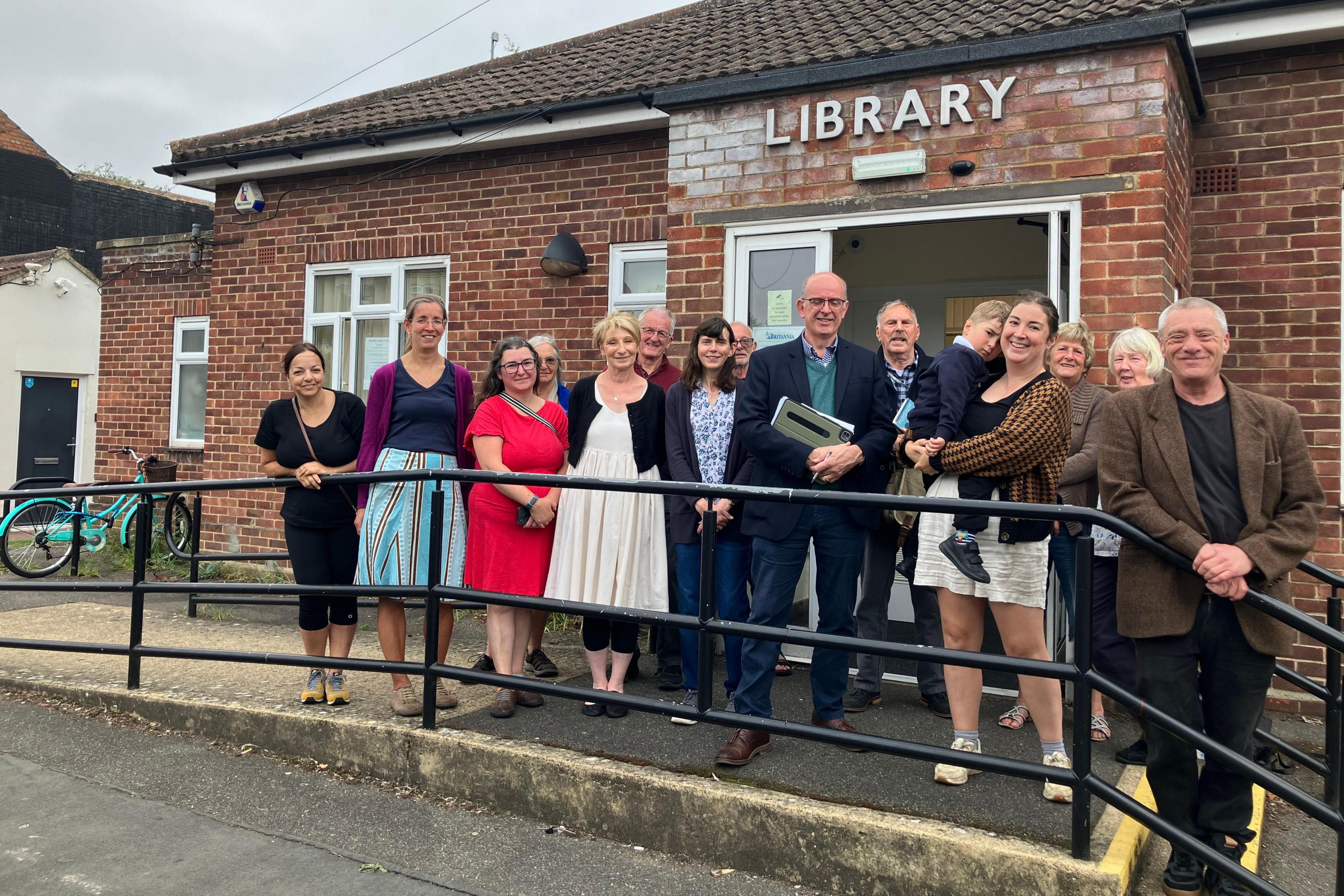 A group of people stand in front of a small red bricked building with the word 'library' written on its front. There are 15 people standing in front of a railing including a woman with a small child