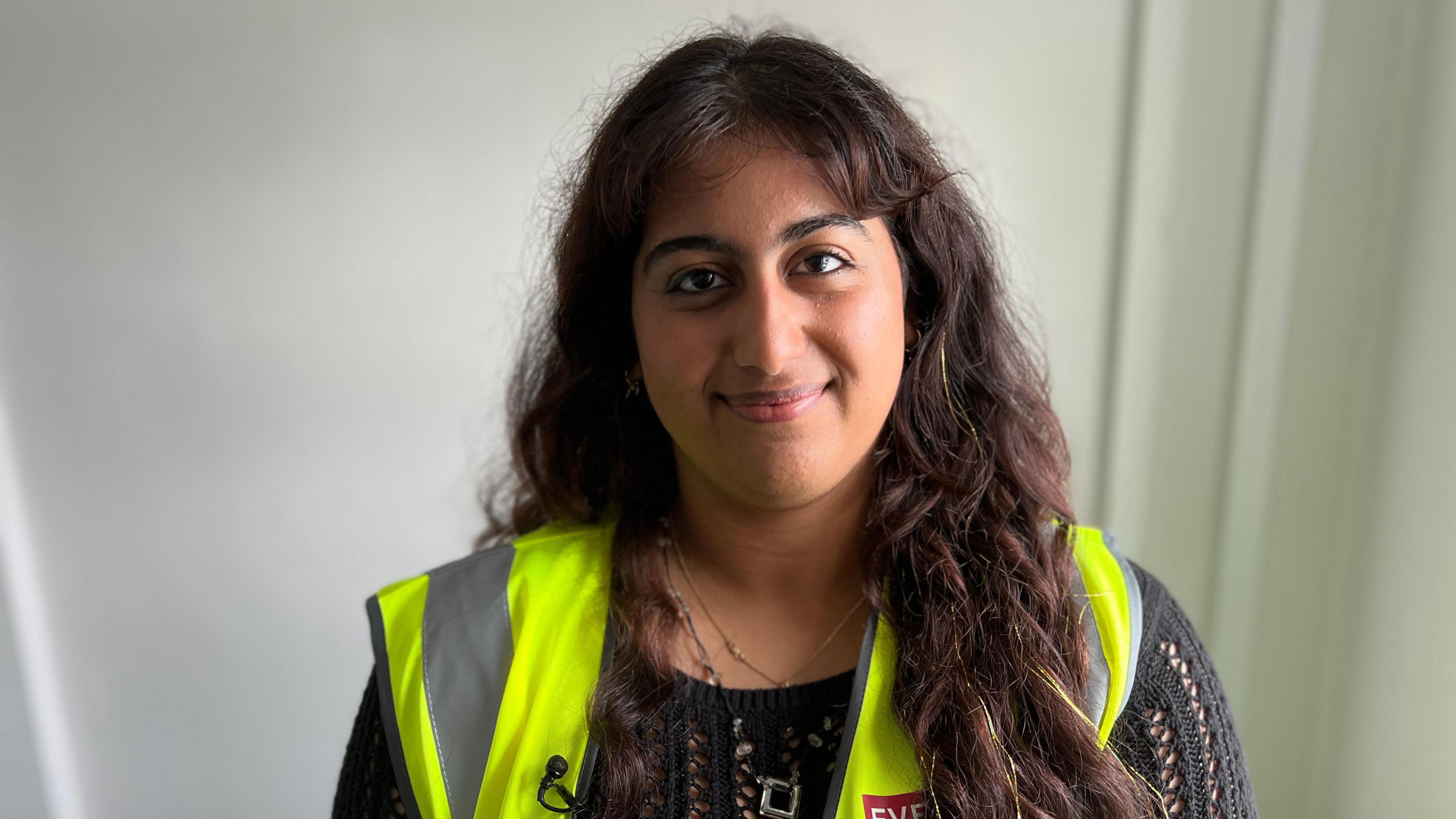 A young woman, with long dark brown hair, smiles as she stands in front of a white wall. She is wearing a black crochet-type jumper and a yellow high visibility vest.