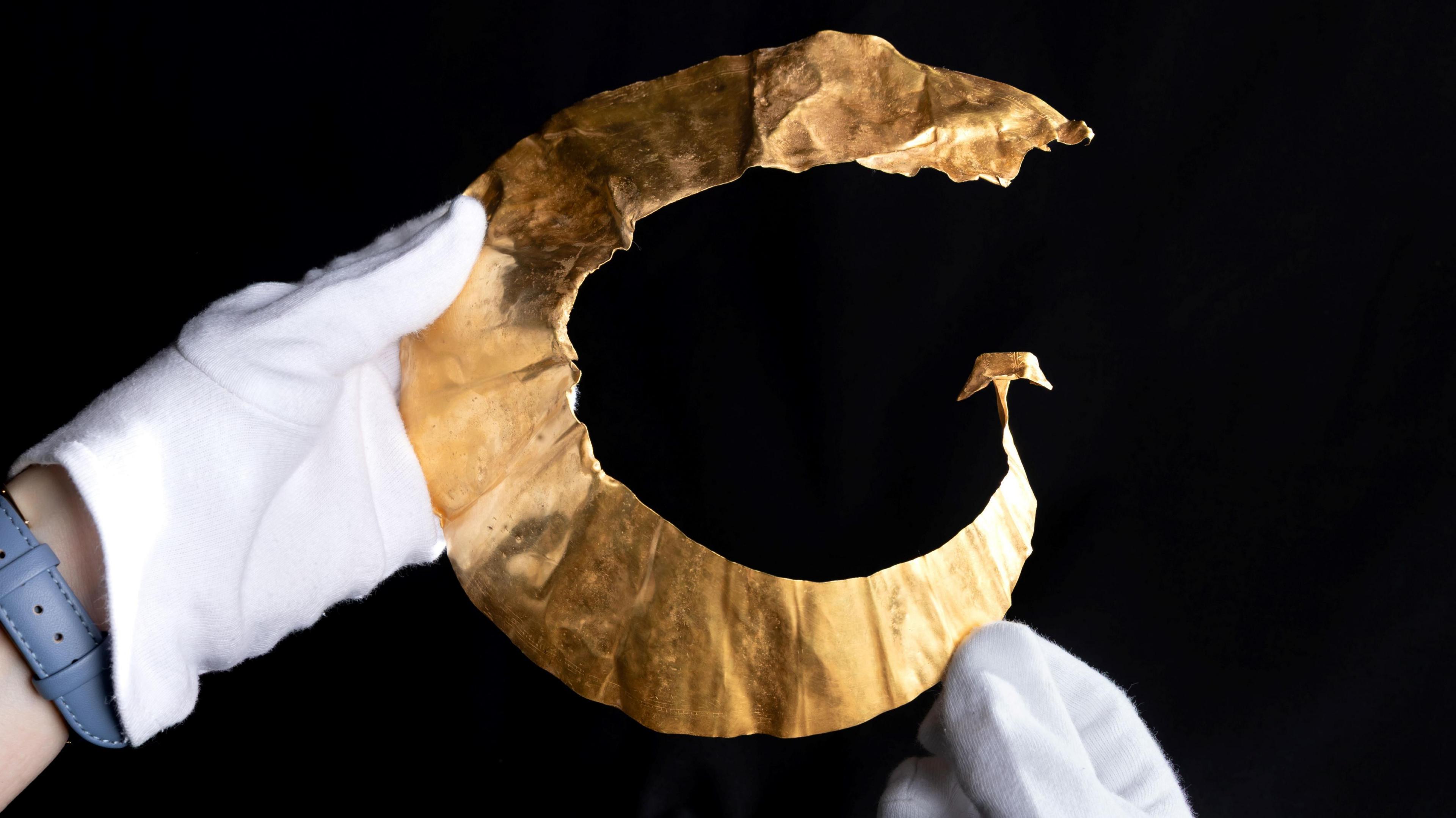 A gold lunula shown up close. It is a crescent shaped gold object handled by someone with white gloves on a black background