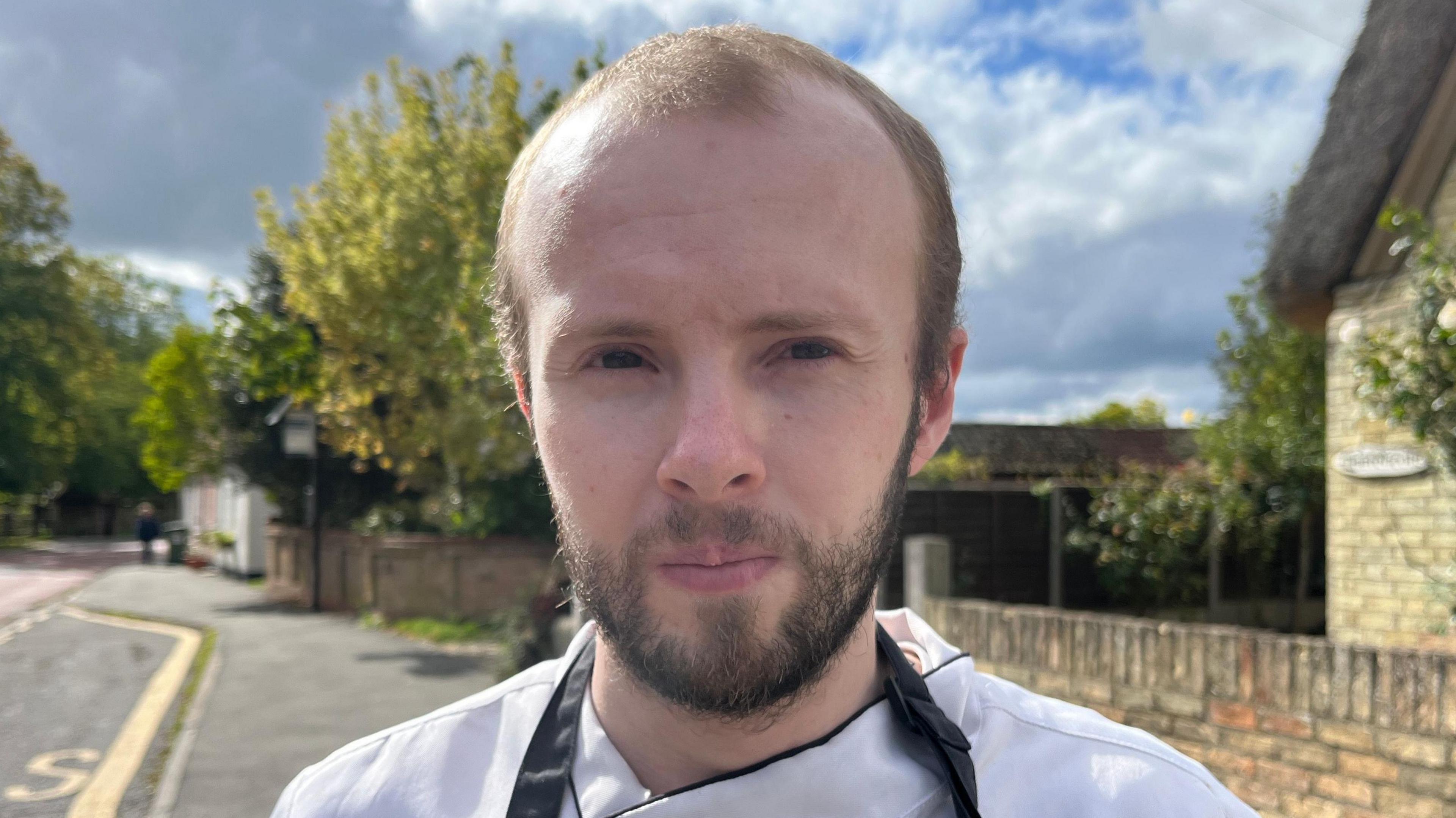 Aaron is standing outside next to a quiet road on a sunny day. He is wearing a chef's uniform - a white shirt with a black apron. He has short dark hair and a beard.