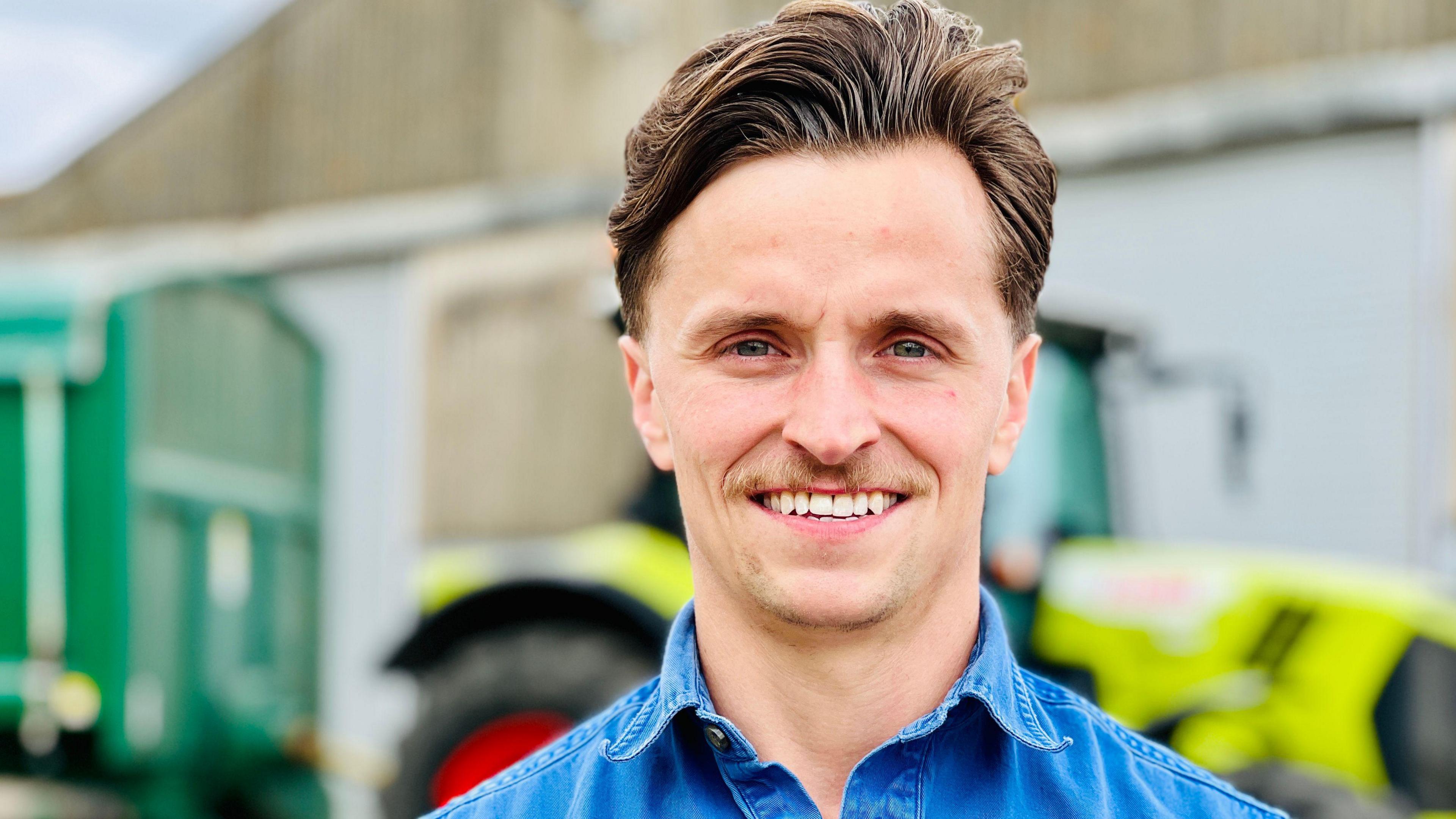 Farmer James Porter smiles as he looks into the camera. He has swept-back brown hair and a thin moustache. He wears a blue denim shirt. 