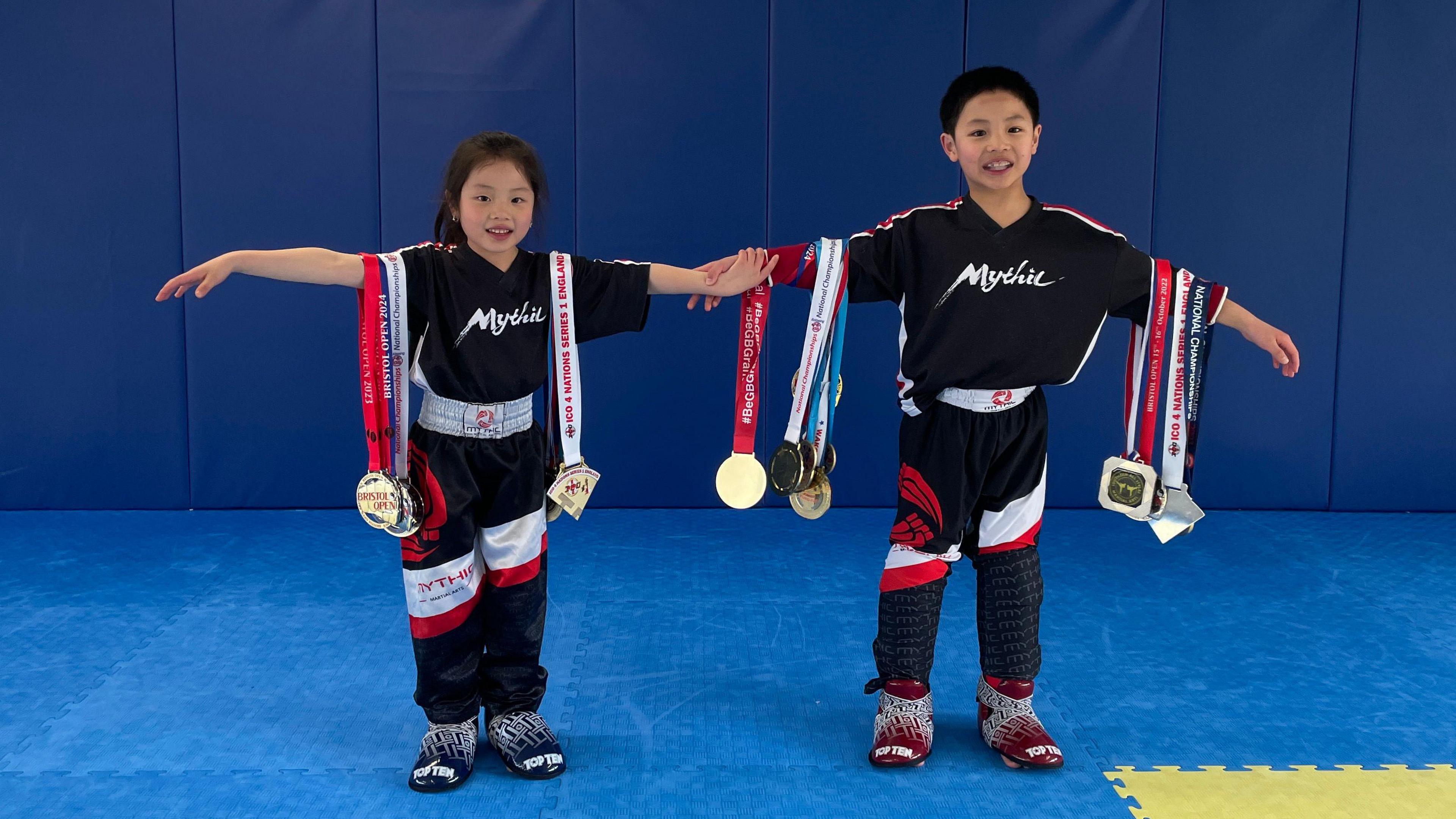 Ivanna and her brother Alex stand side by side with their arms out. They have all of their medals, at least ten, hanging from their arms. 