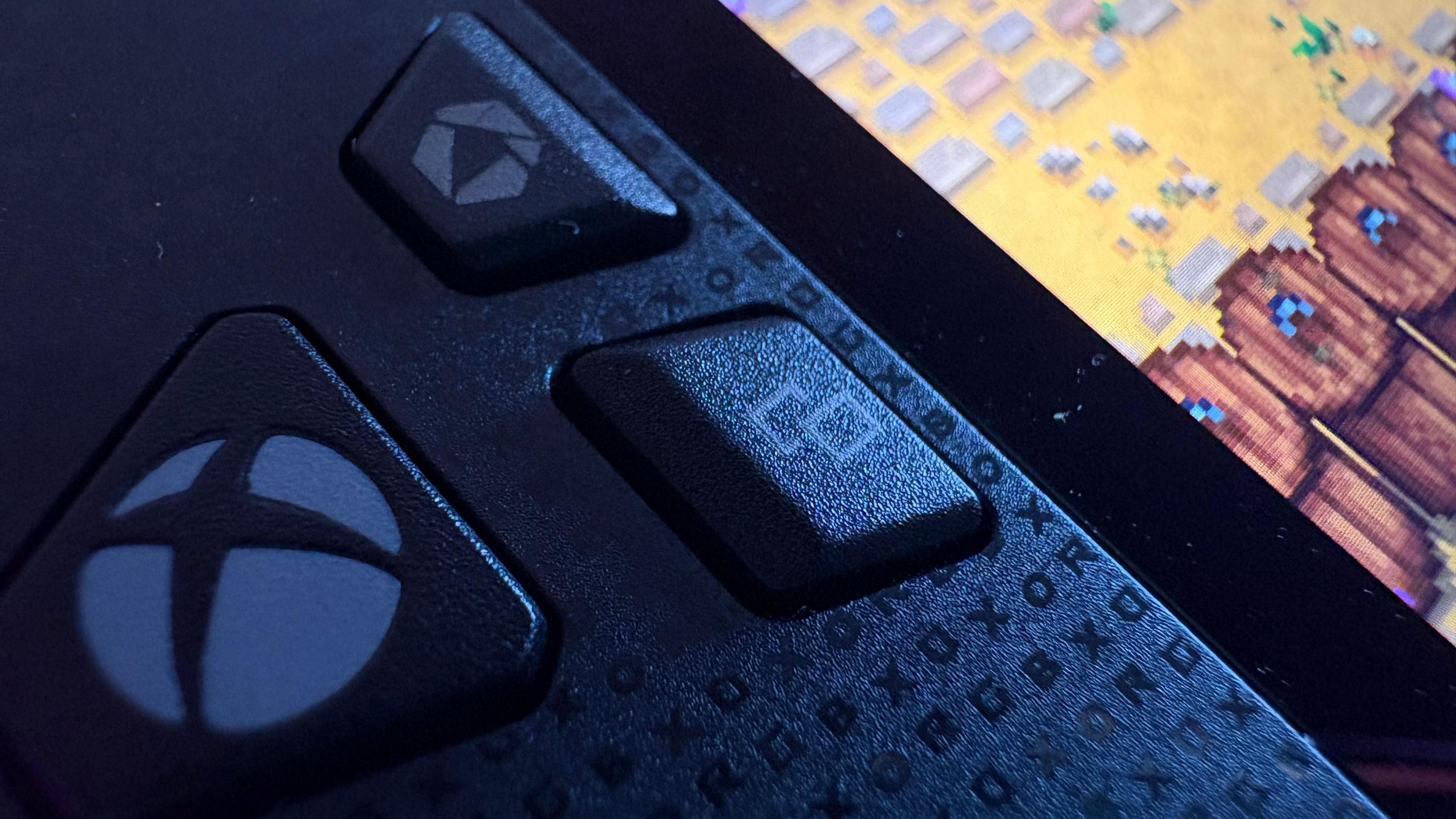 A close-up of the Xbox logo on the handheld. It is an Xbox logo. Beside it are two other buttons. We can only see a portion of the screen, but it has bright pixel graphics with what appears to be several barrels. It is from Stardew Valley.