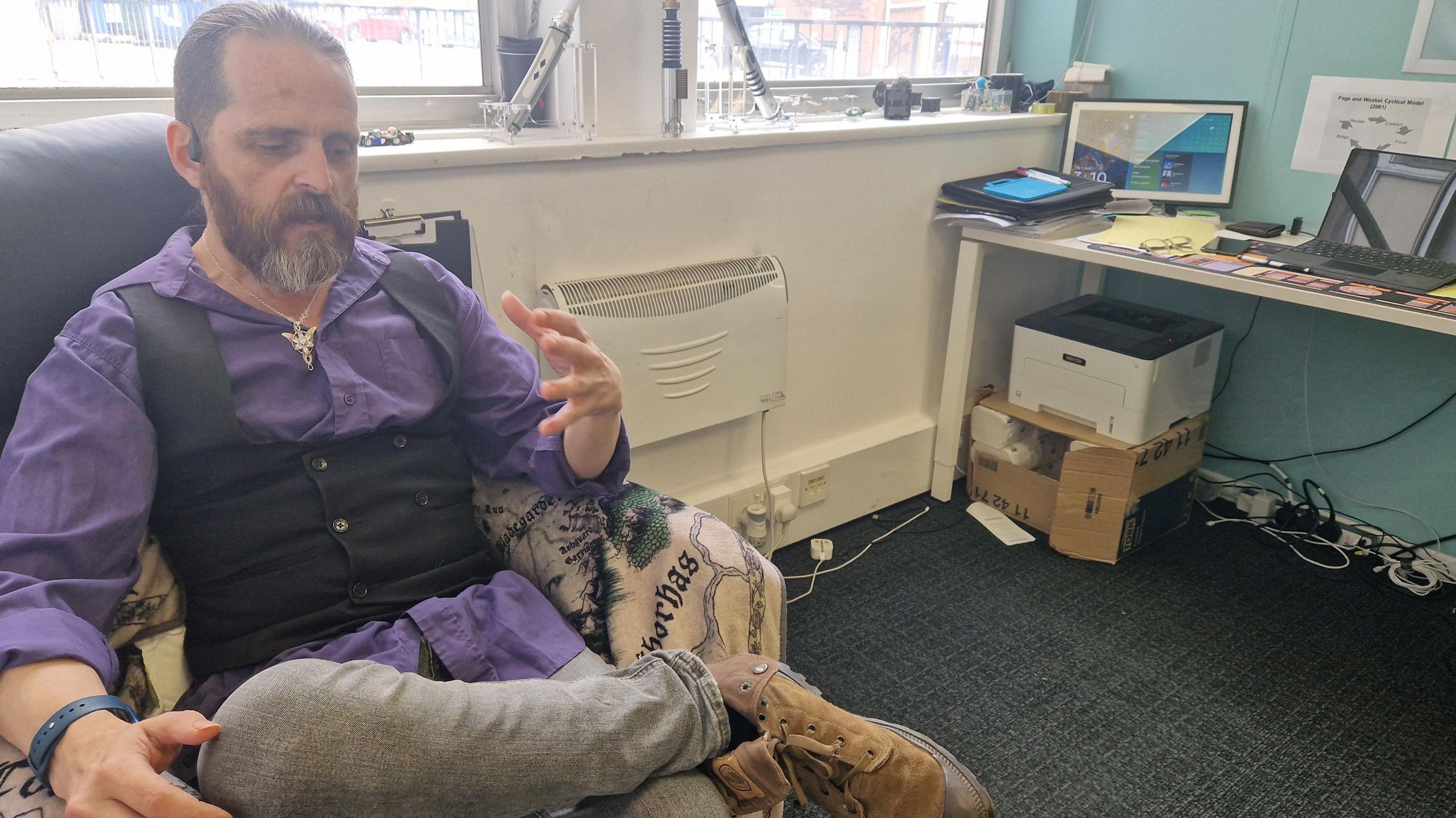 Rob has dark hair tied back in a pony tail and a beard. He is wearing a purple shirt which is open at the collar, a black waistcoat and a large silver pendant necklace. He is sitting in his office and chatting. The chair he is sat in is to the left of the image. On the right is a desk with a laptop and various paperwork. Underneath is a printer and a box, along with an extension cord with various plugs and wires. There is a window behind him and on that wall is a heater mounted. 