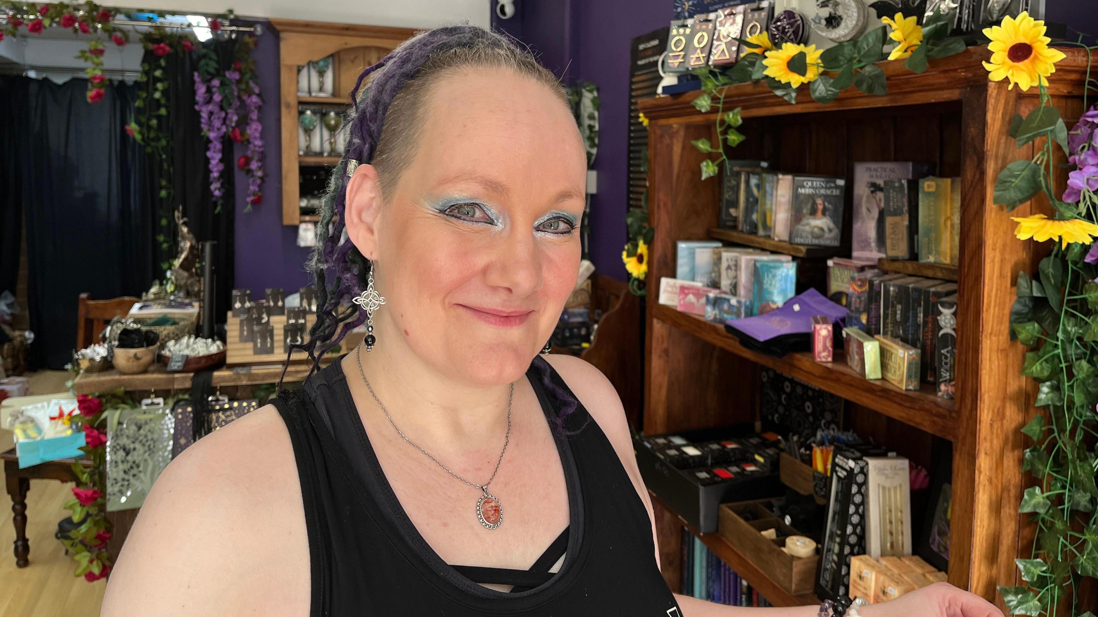 Ms Archer standing in her shop. She is wearing blue eyeshadow and large silver jewellery. In the background you can see flowers, books, jewellery, crystals and charms