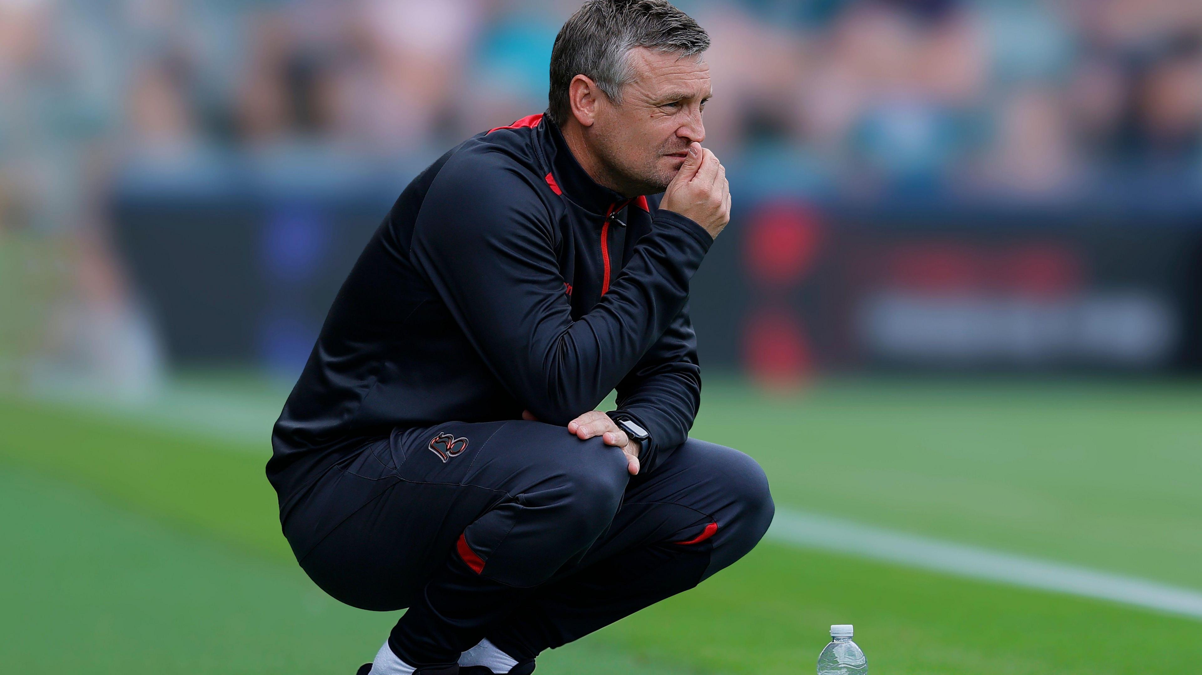 Bristol City manager Michael Skubala, in a dark blue tracksuit with red trim, squats at pitchside, with his hand to his mouth watching his team in action.
