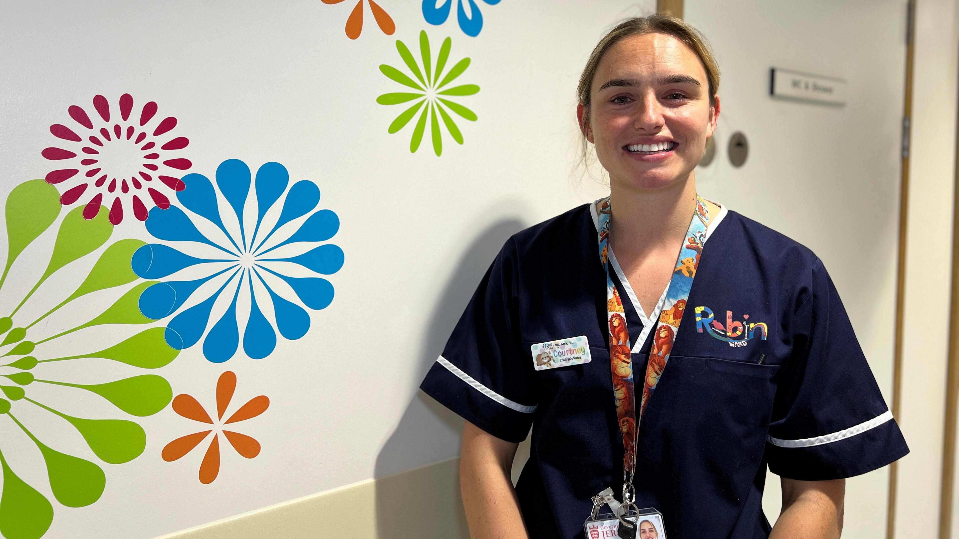 Courtney smiles at the camera and she is wearing her nurse uniform. She's stood next to a wall in the hospital corridoor with some art designs on it. Her dark blond hair is tied into a ponytail.  