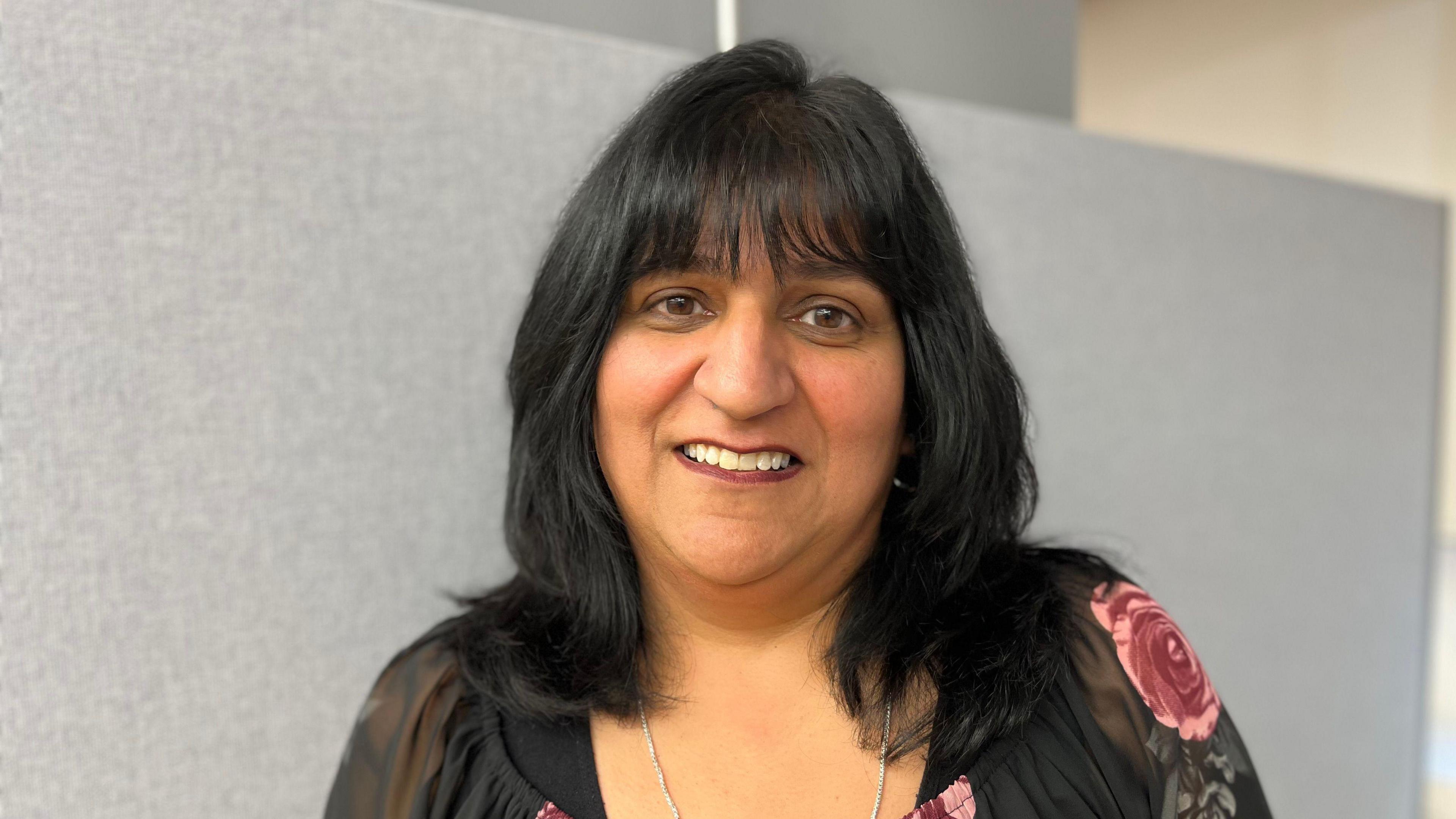 A headshot of Rooprit Gill wearing a black and pink floral blouse. There is a grey background.