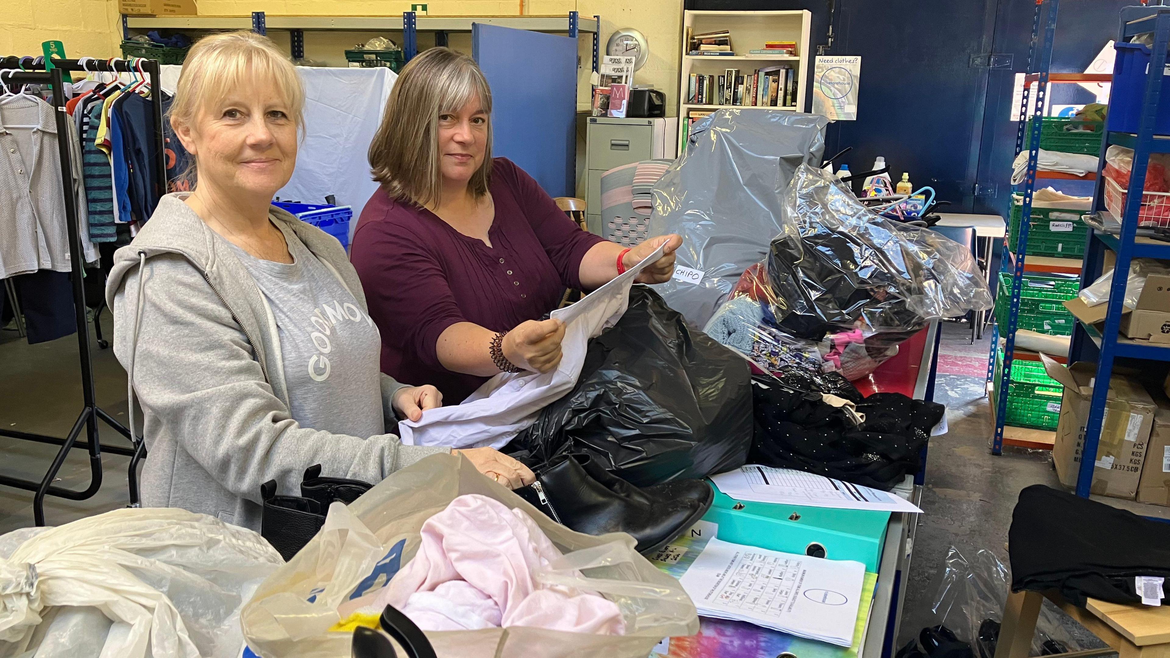 A picture of two women, one with blonde hair, the other with brown hair, rummaging through bags of clothes.