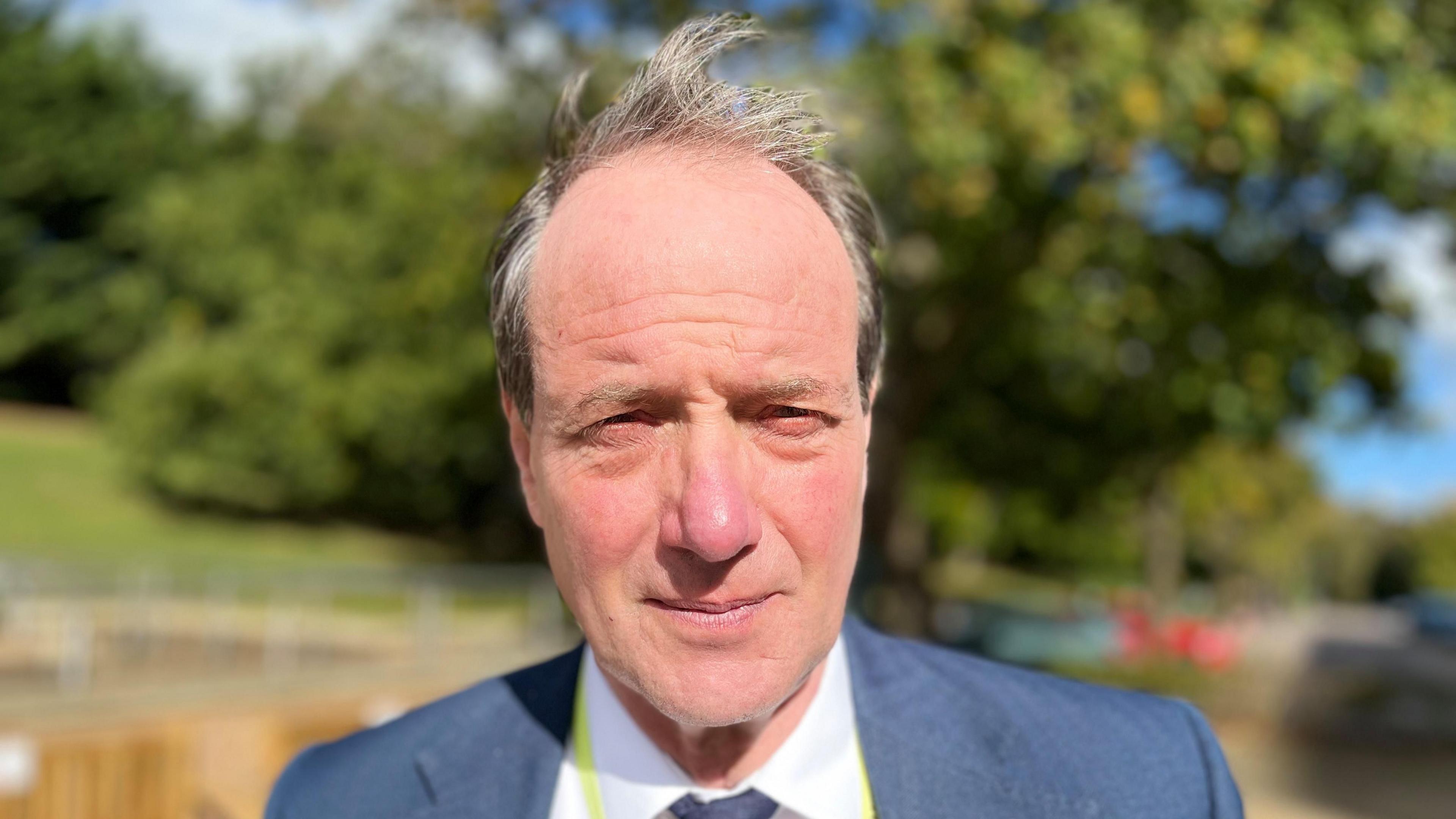 Andrew Jamieson, deputy leader of Norfolk County Council, looking into the camera outside County Hall in Norwich. He is wearing a blue jack, white shirt and blue tie. There is greenery in the background.