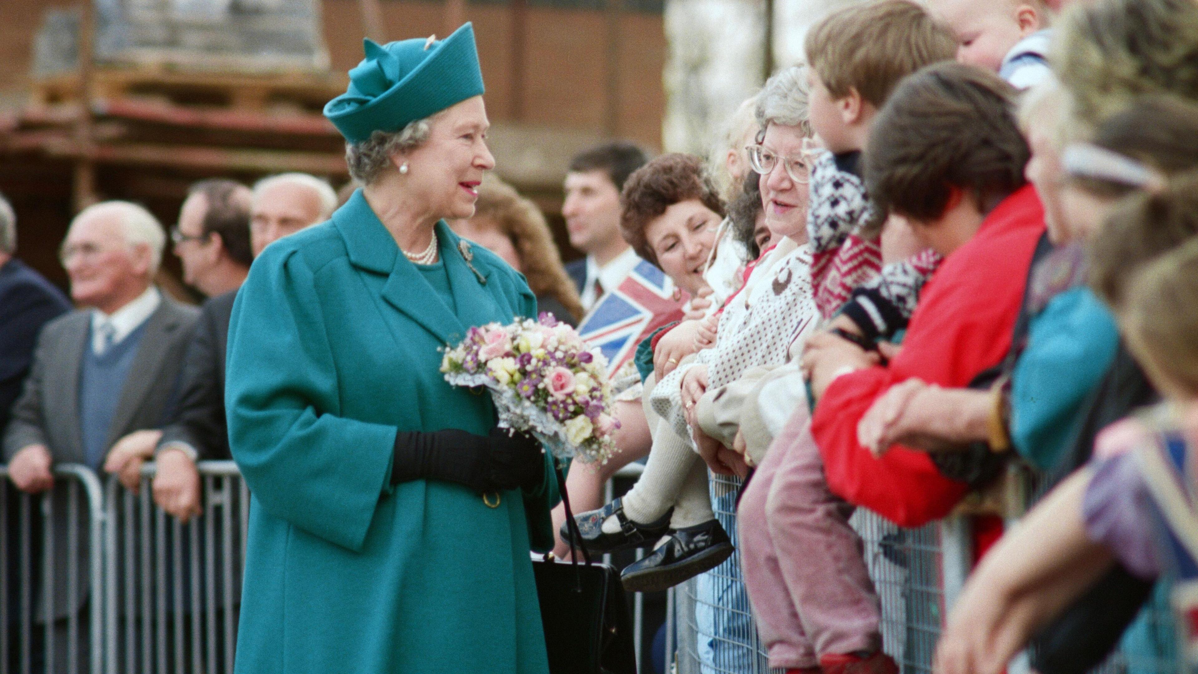 Queen Elizabeth II wearing a green/blue coat and hat. She's talking to people in the crowd and is holding pink, white and purple flowers.