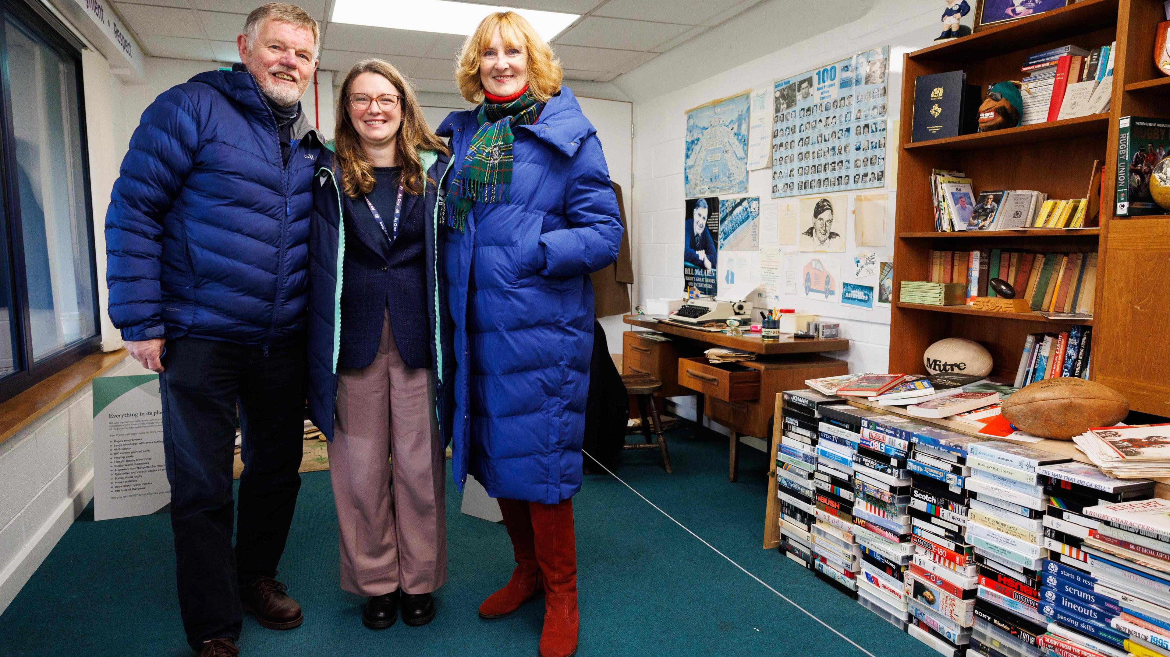 Three people - two women and a man - stand in a recreation of Bill McLaren's study at Murrayfield with books, maps and rugby balls and more memorabilia around them