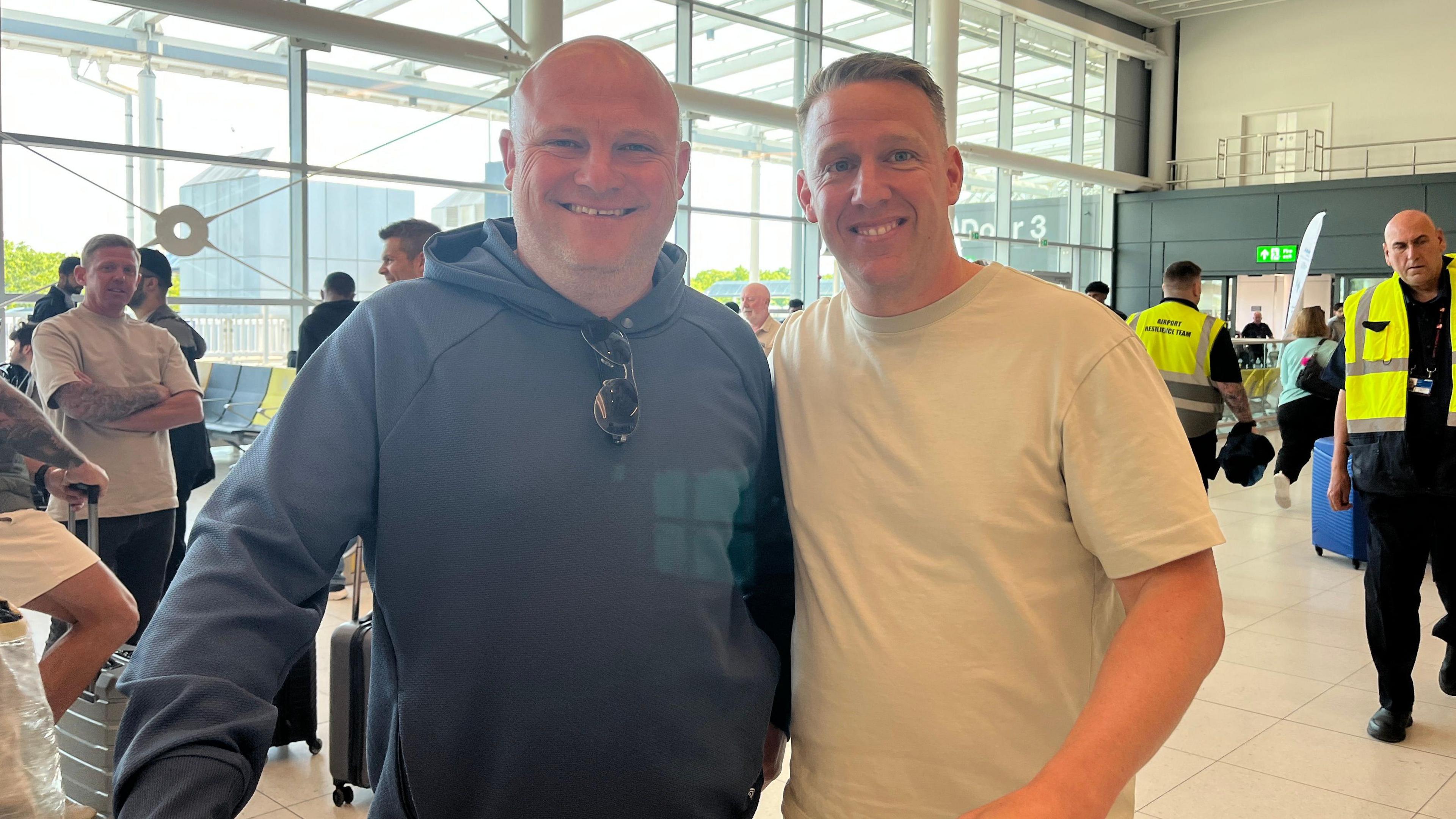 Bob Firth and Nathan Hall stood together smiling at Manchester Airport. Bob has a bald head and us wearing a grey designer hoodie. Nathan has short brown hair and is wearing a white T-Shirt