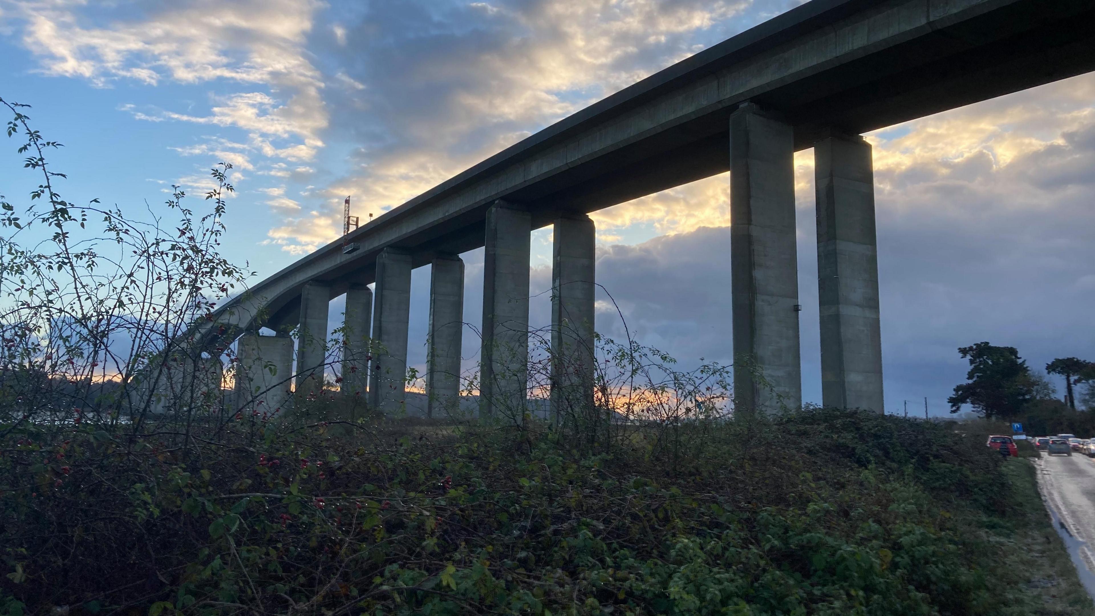 A view of the Orwell Bridge during a sunset where the sky is blue but also full of white and grey clouds. There's a burst of orange on the horizon.