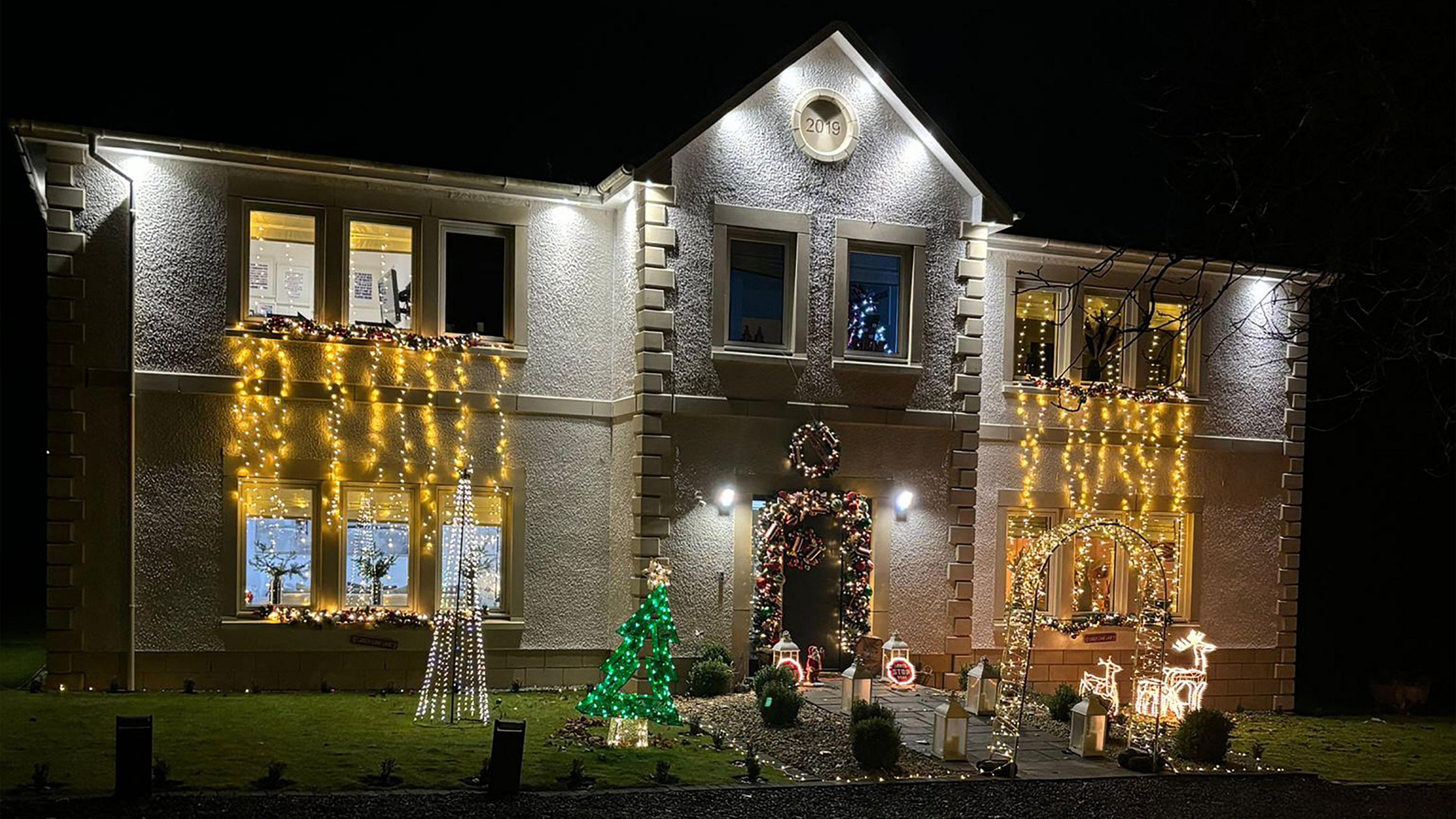 A large, two storey house adorned with light up Christmas decorations. Long yellow fairy lights dangle from the top floor windows. The front garden is full of light up ornaments including a Christmas tree, a reindeer, lanterns and an archway. A Christmas wreath hangs above the door and the door is bordered by Christmas foliage. 