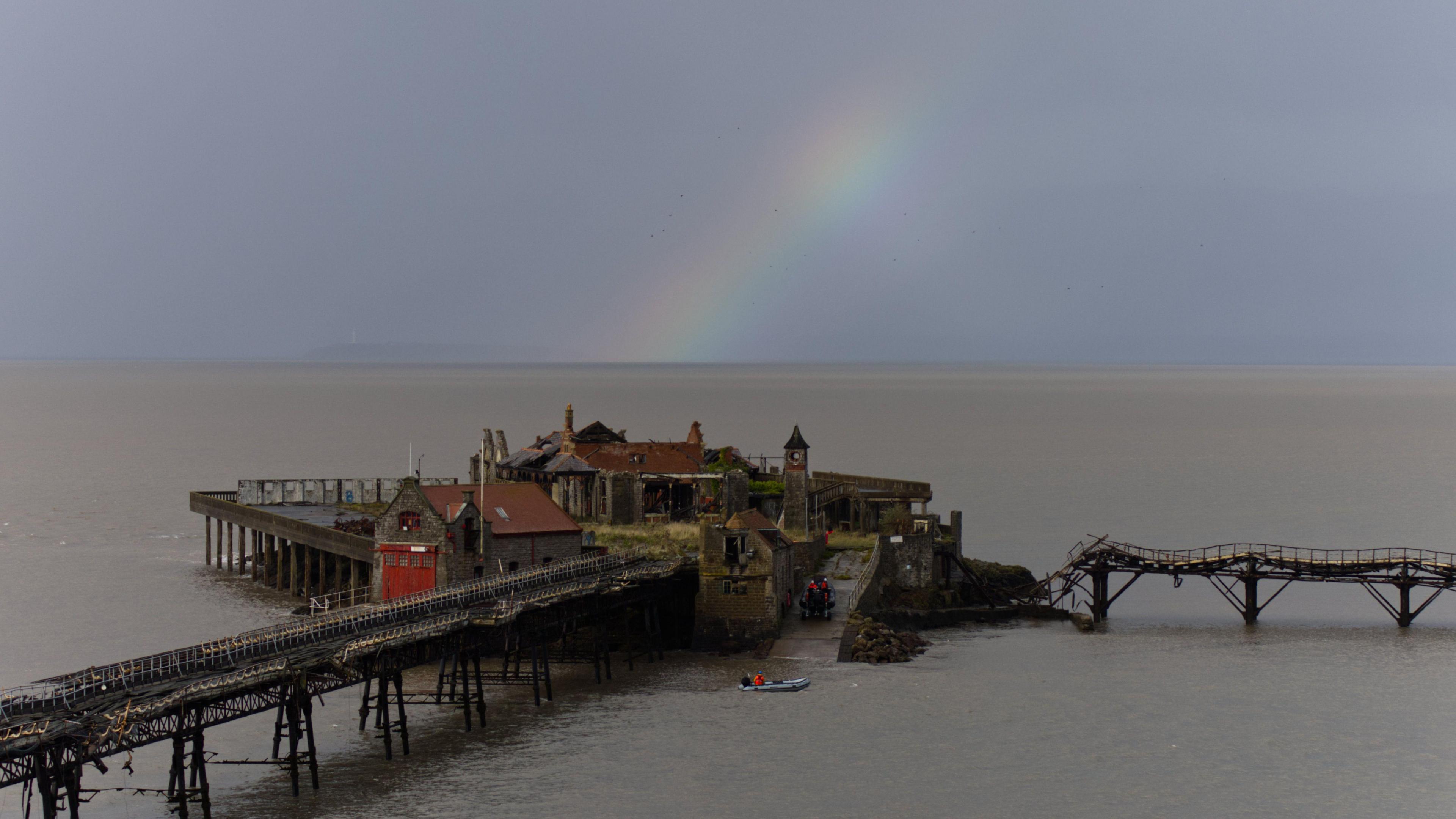 A pier that is falling apart is seen under a rainbow and grey sky.