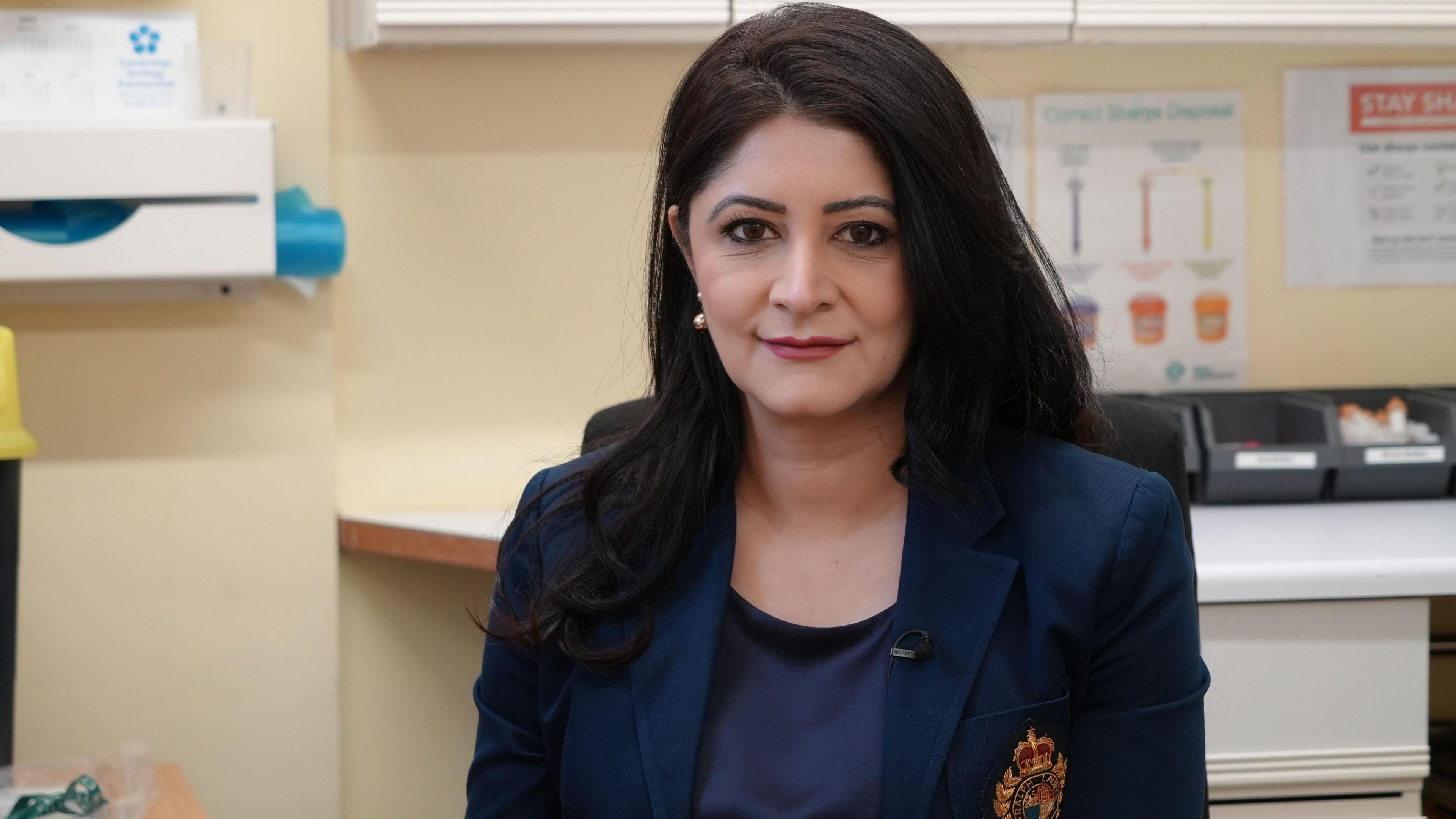 Dr Shabina Asad Qayyum smiles at the camera while sitting in an office. She is wearing a navy blue top and a navy blue blazer with an orange logo. She has long black hair. 