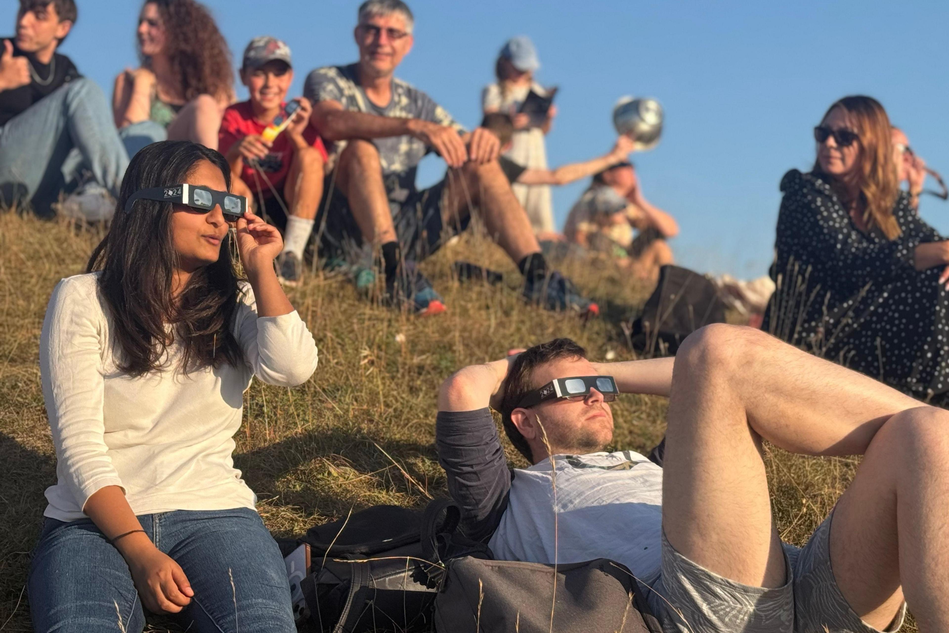 People wearing eclipse glasses sit and lie on a grassy slope as they watch a partial solar eclipse. Somebody is holding a colander in the background