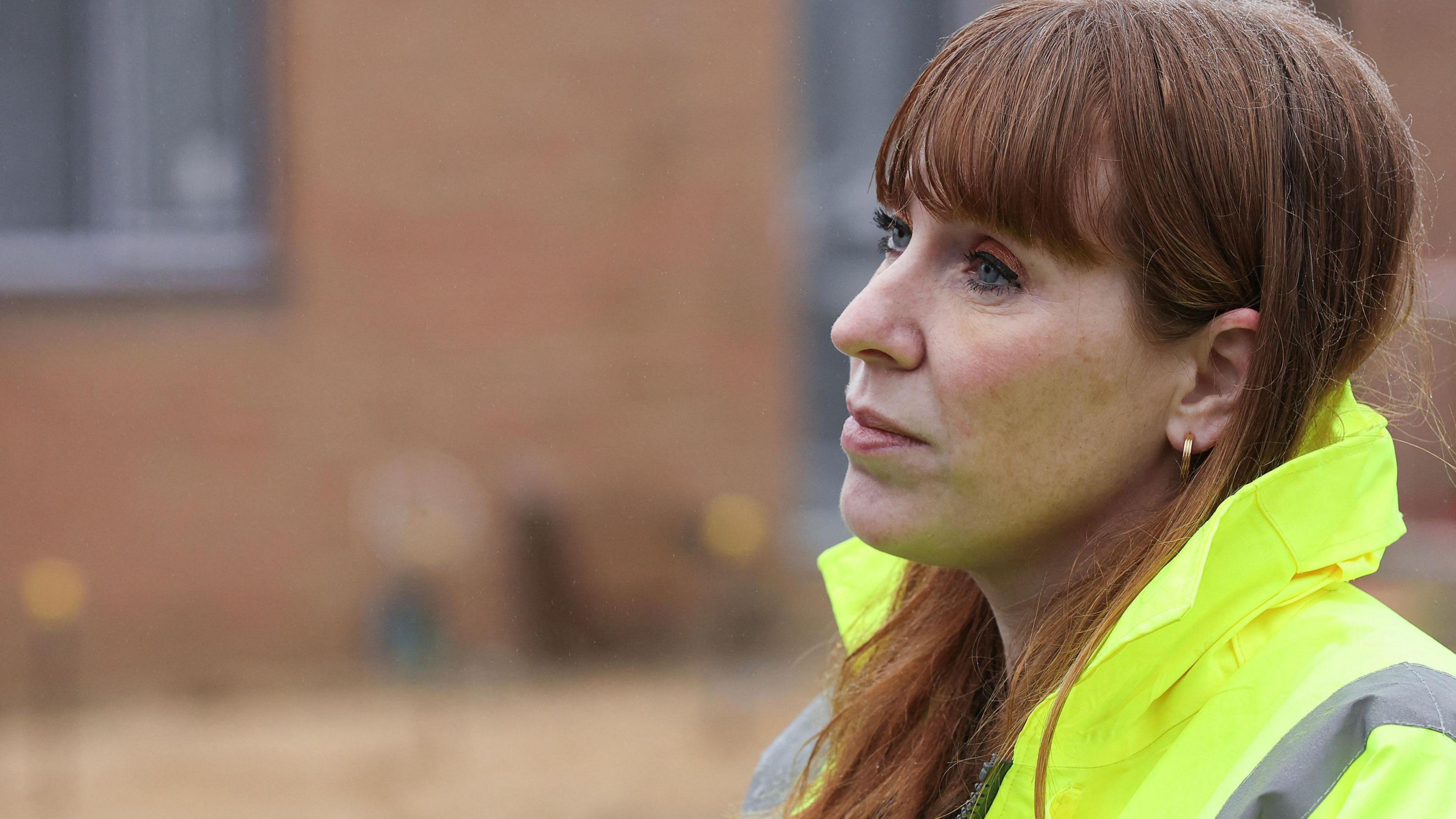 Housing Secretary and Deputy Prime Minister Angela Rayner, with long auburn hair and gold hoop earrings, wears a yellow hi-vis jacket at a construction site.