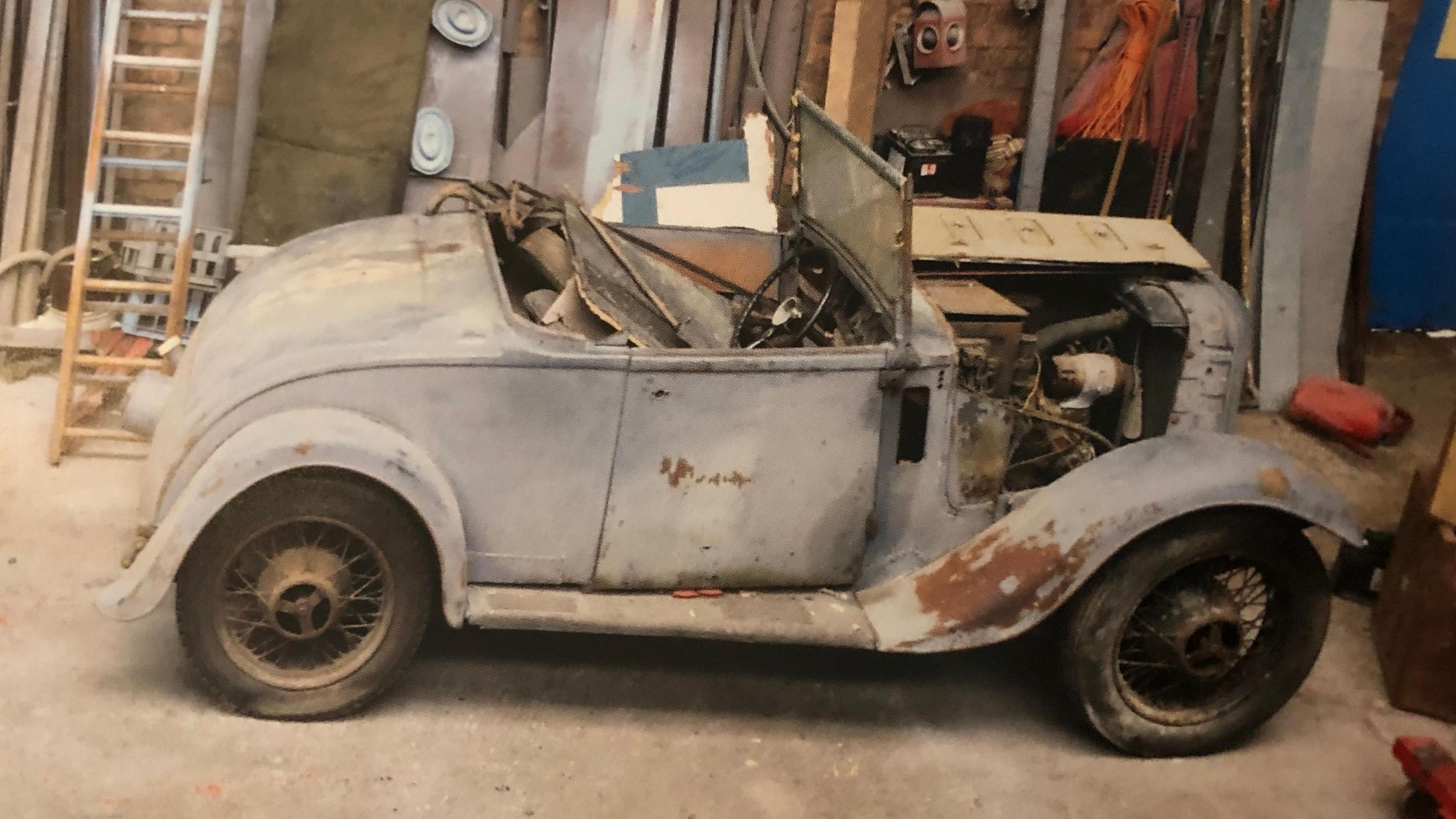 An old and damaged white-coloured classic car parked in a workshop. A ladder is leaning up against a wall on the left. Against the wall behind the car are some tools and clothing items.