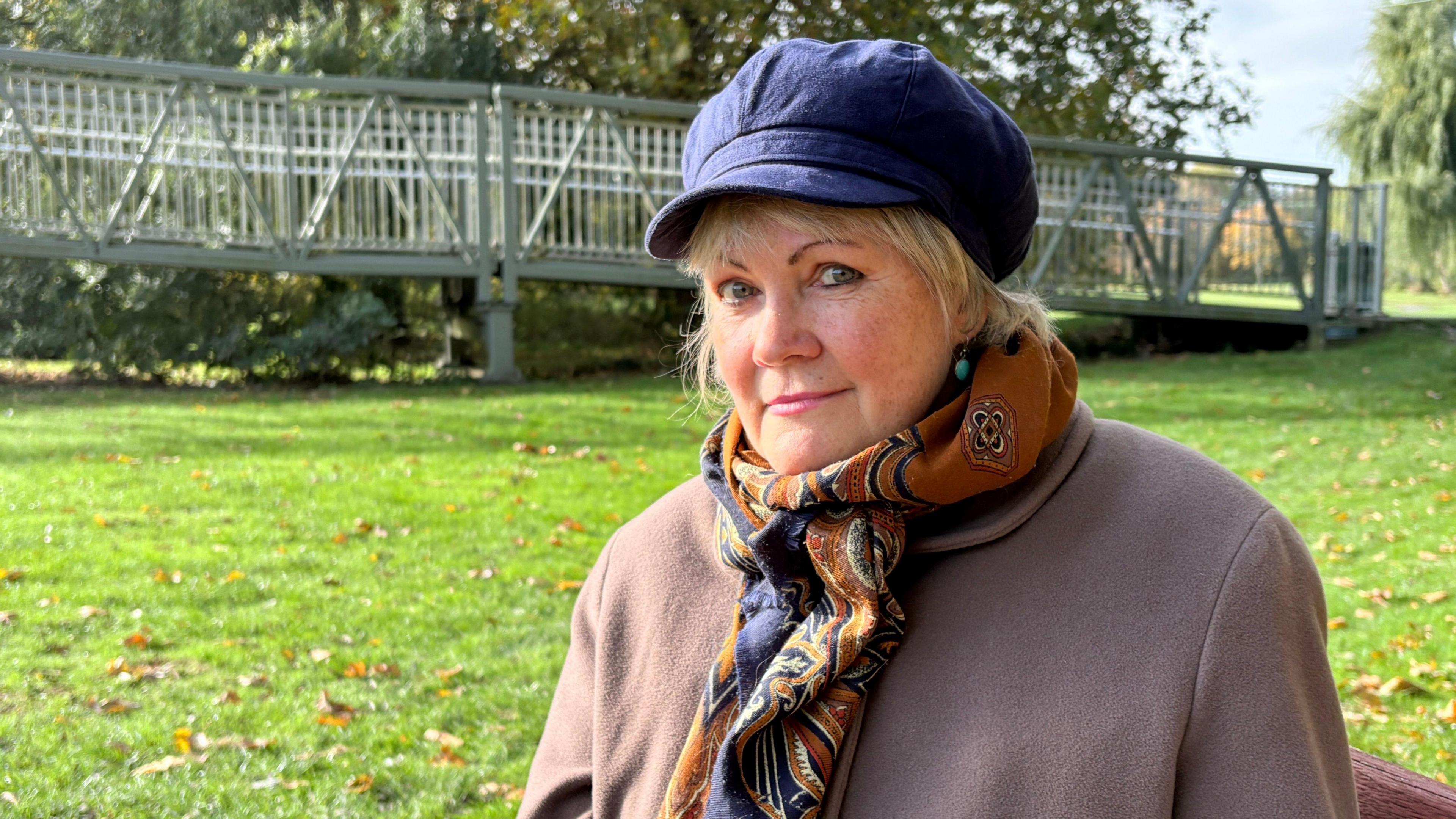 Christine Green, sitting outside in a park on a bright, sunny day. She is wearing a peaked blue cap, from which her blonde hair is just emerging, a rust and navy blue scarf, and a heavy-looking light brown coat. Her blue eyes are looking down the camera. She is seated on a bench, whose back can just be seen on the bottom right corner of the image. Otherwise, there is brightly lit grass behind her, dappled with fallen leaves. A pedestrian bridge is in the further distance, along with some trees, which are out of focus.