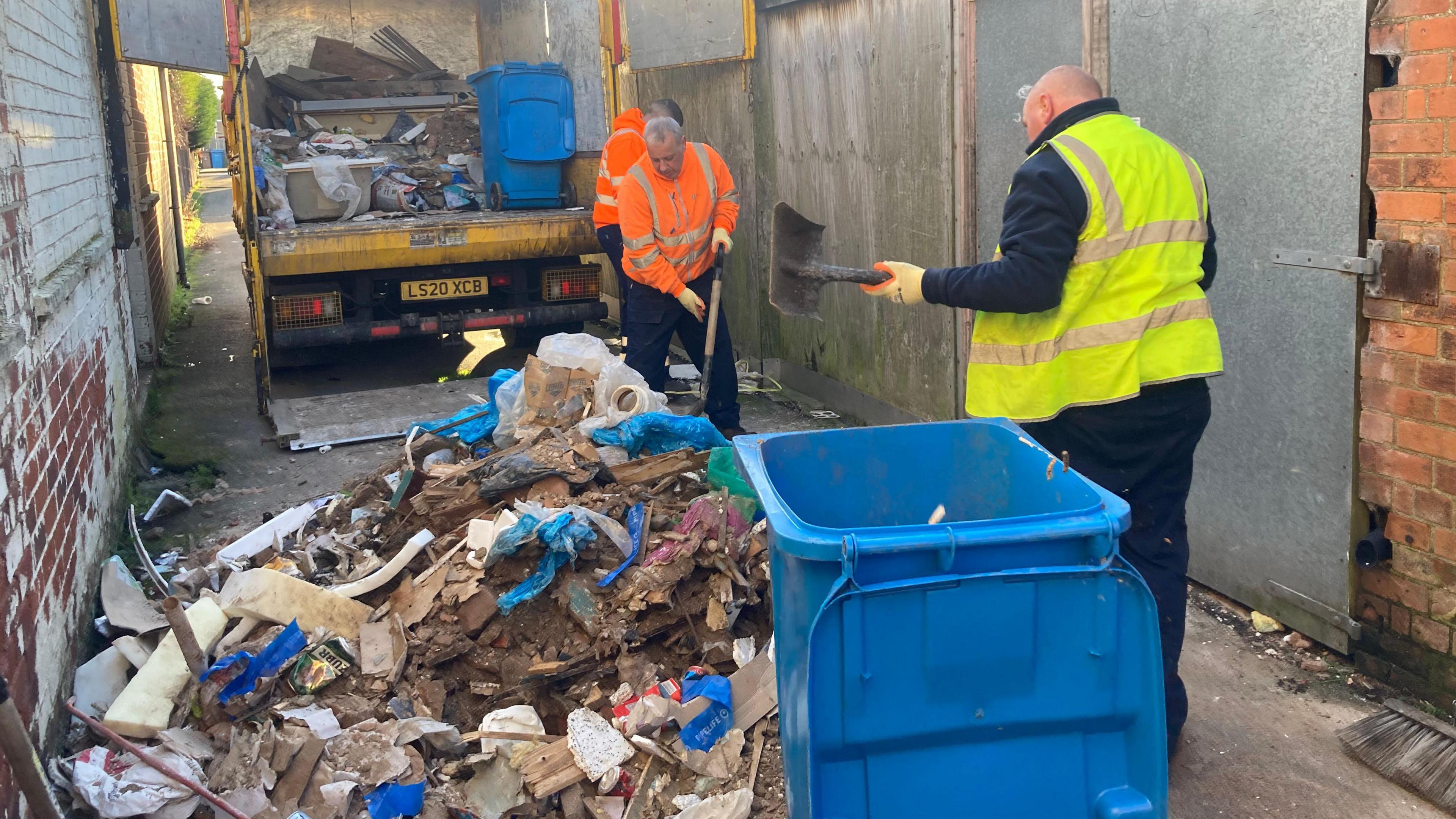 A large pile of rubbish dumped on the floor in an alleyway. Three men, two in orange hi-vis vests and one in a yellow hi-vis vest scooping up the waste with metal shovels. A yellow lorry carrying rubbish and a blue bin is parked with its rear facing the camera. There is an open blue bin in the forefront of the picture. 