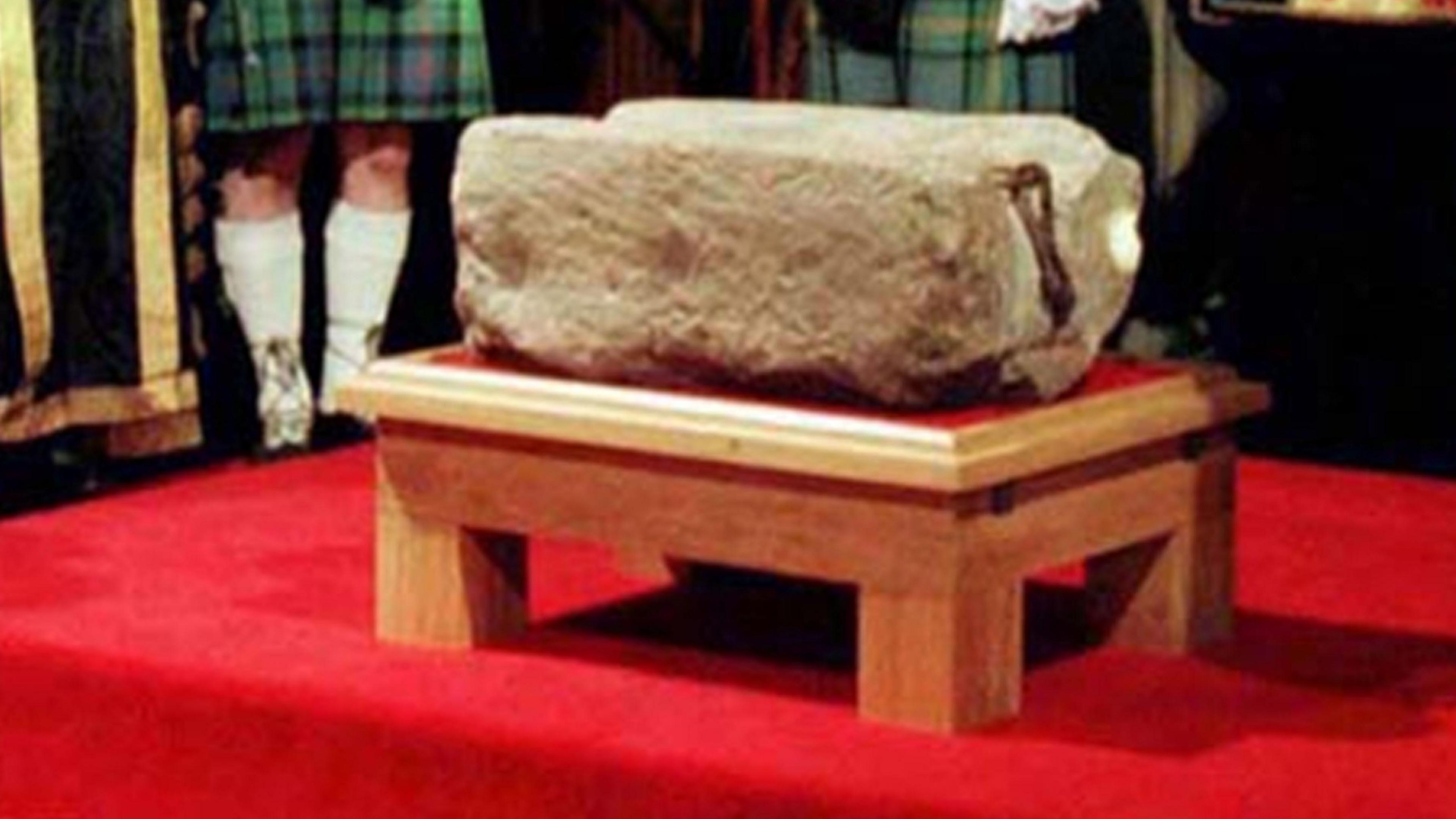 The stone of destiny sitting on a wooden plinth with a red top. The stone is rectangular in shape, with a rusted hook at one end. It is on a small plinth on a red carpet. The legs of two men in green kilts can be seen in the background.