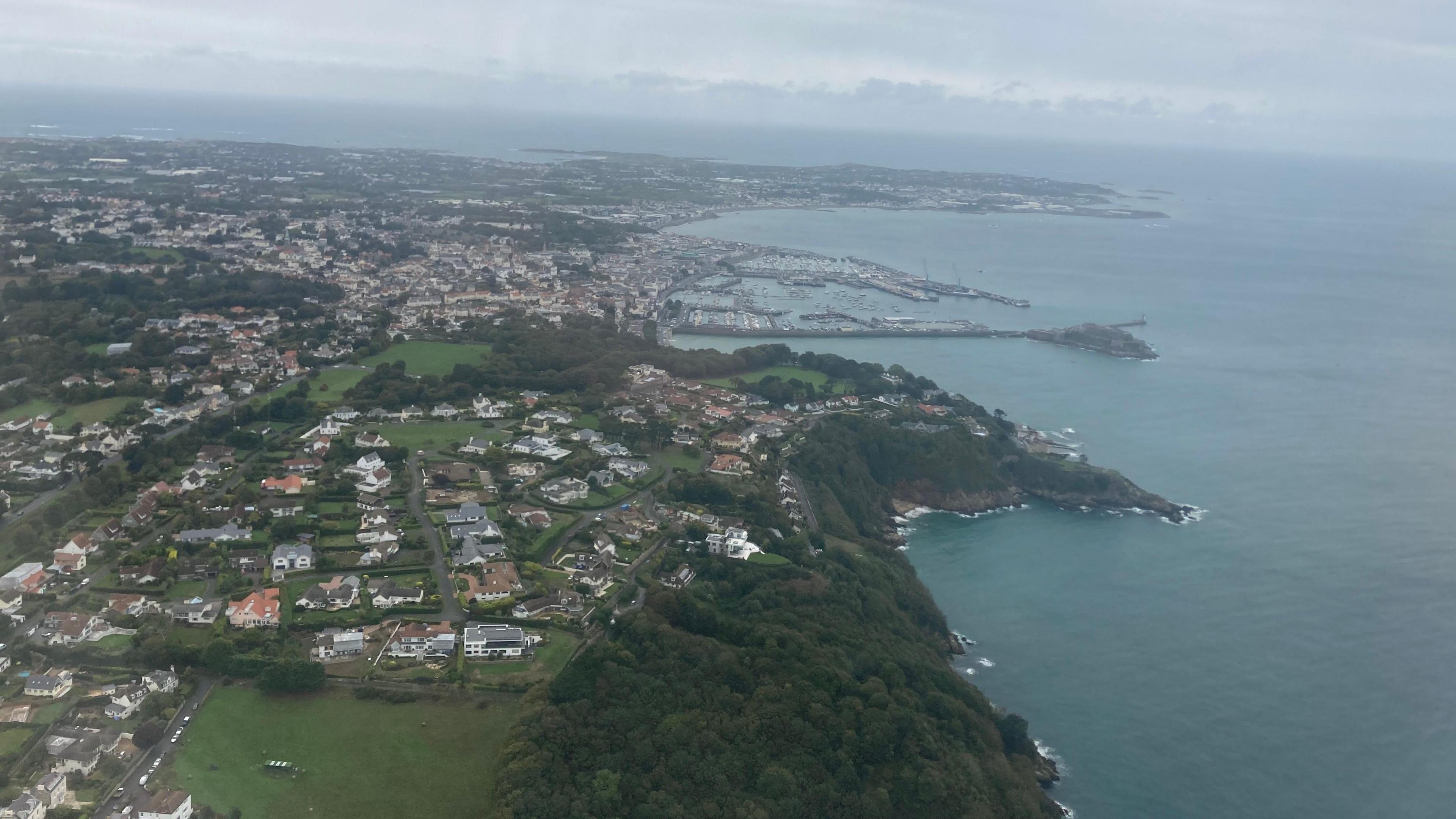 Arial view of Guernsey. The sea is a dark blue and the sky is grey and full of clouds.