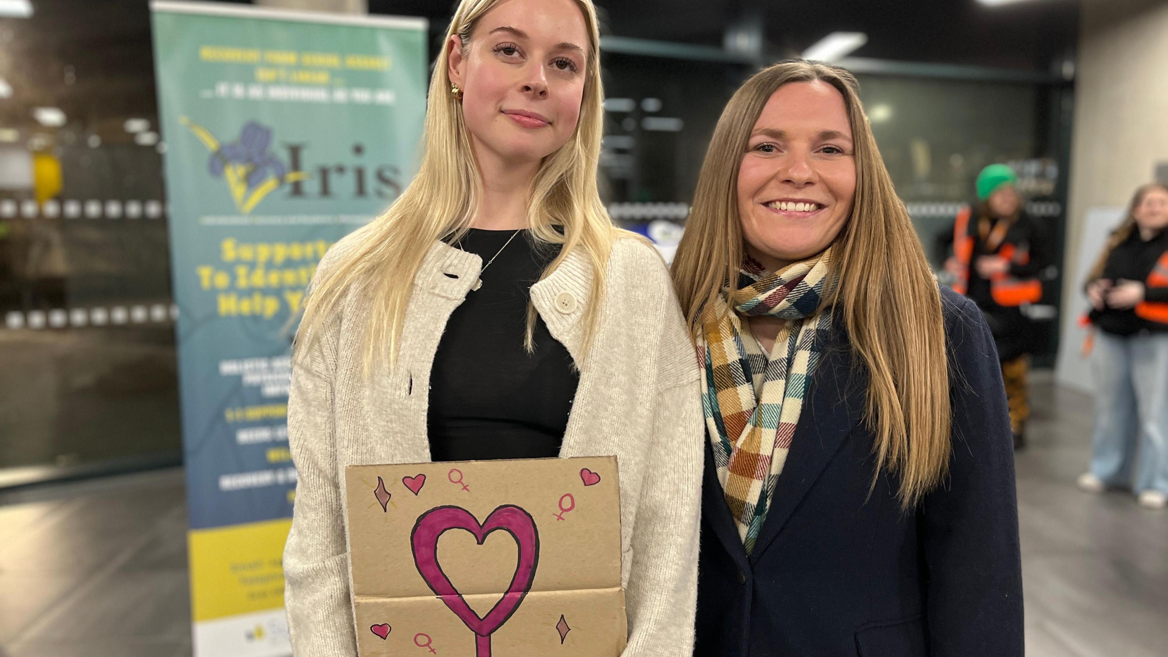 Two women stand, one is holding a sign with a heart logo on it.