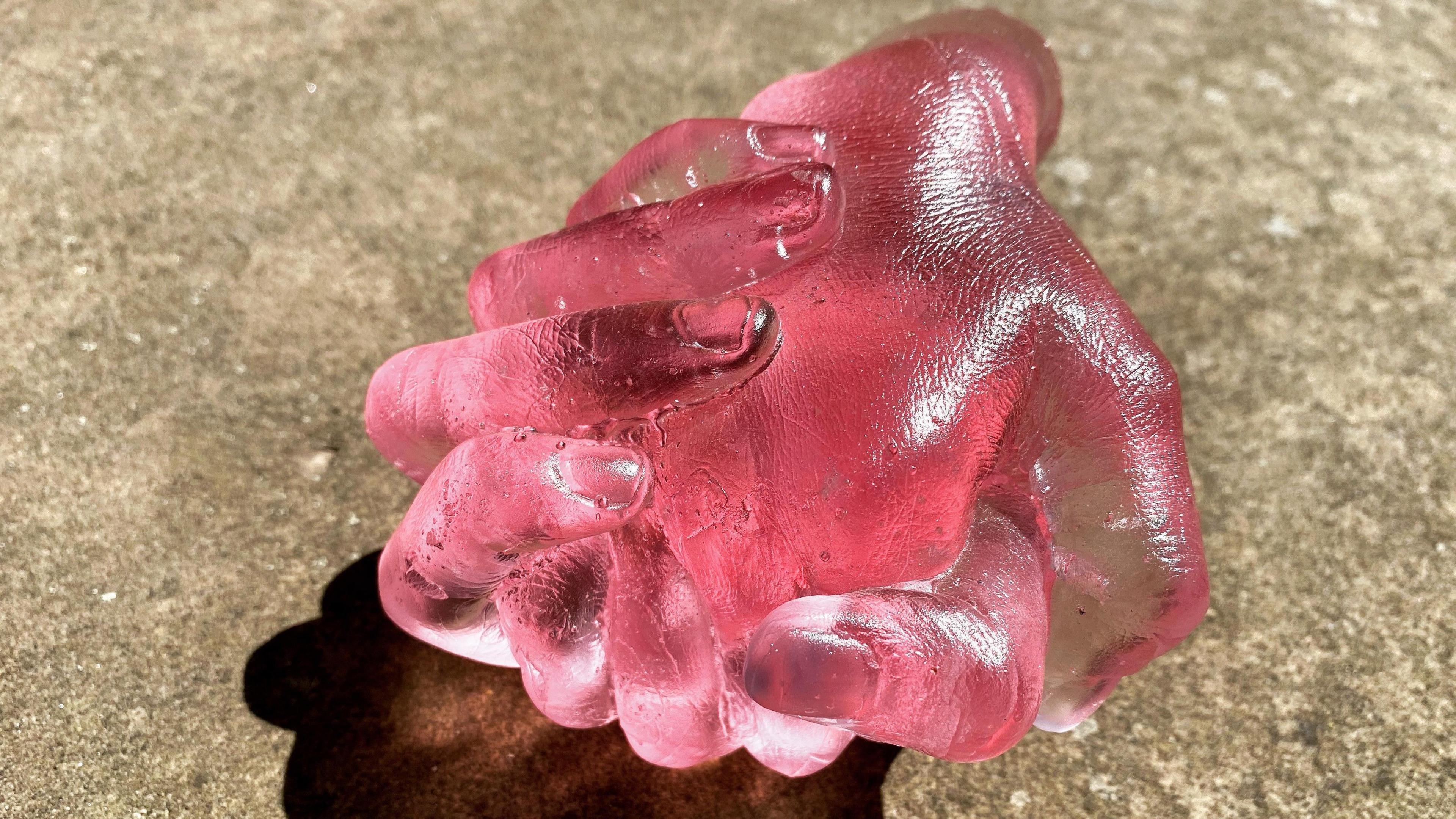 Photo of pink glass sculpture which depicts two hands clasping each other. It is very realistic looking showing the fine lines of "skin" in intricate detail.