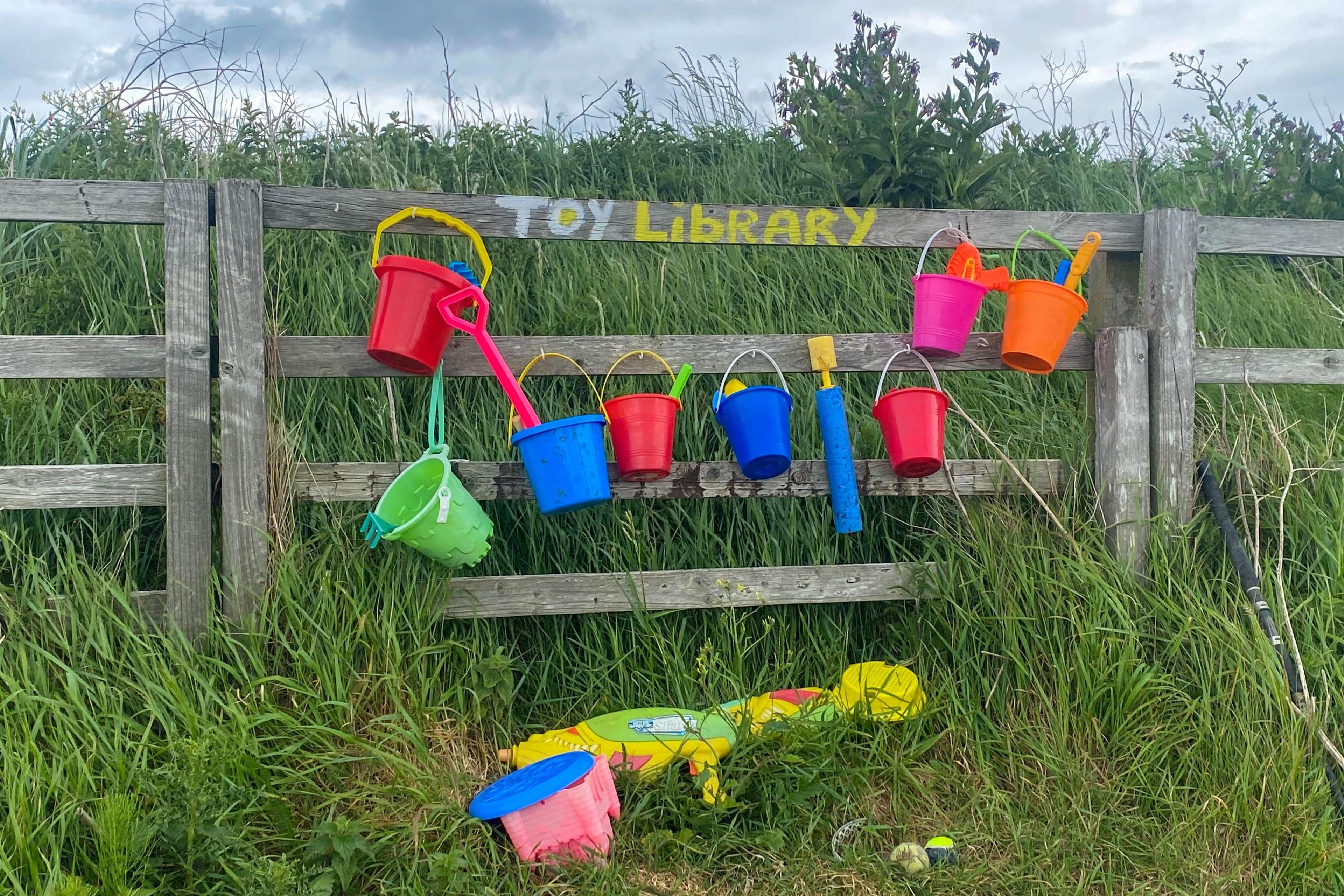 Bright toy buckets and spades in green, red and blue, on a wooden fence, amid grass, with the words 'toy library' painted on the fence.