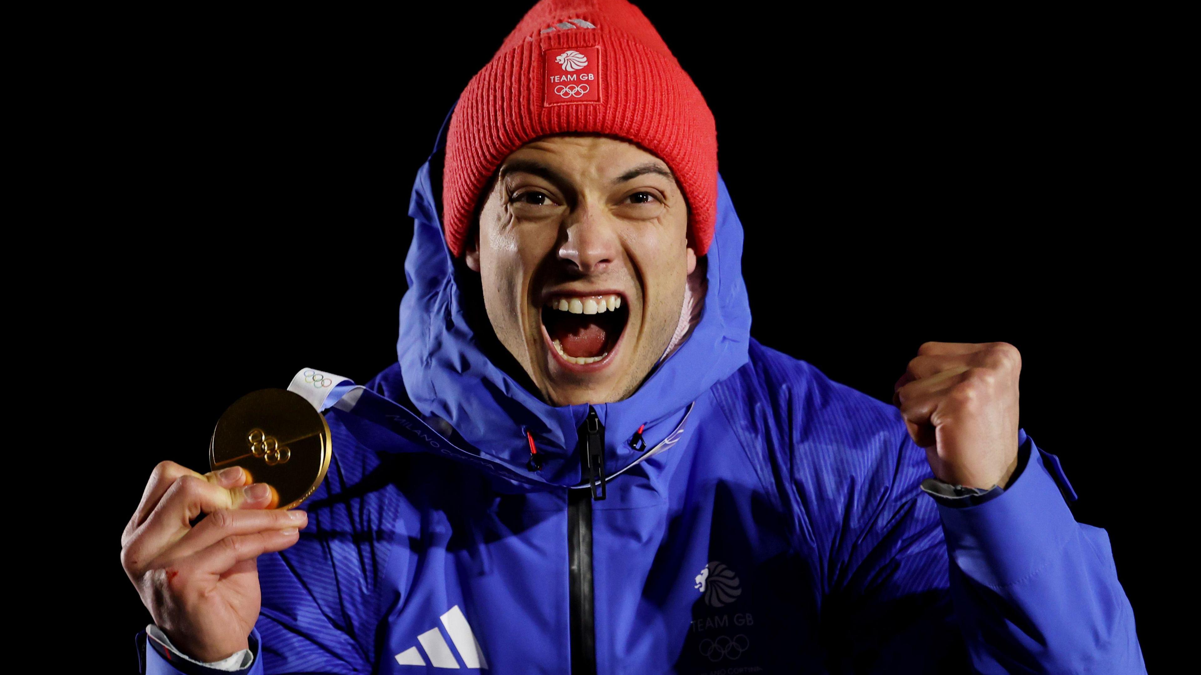 Matt Weston wears a red hat and blue coat. He is holding his gold medal up with one hand and the other hand is held in a fist in a celebratory manner. His mouth is open as if he is screaming in celebration. 