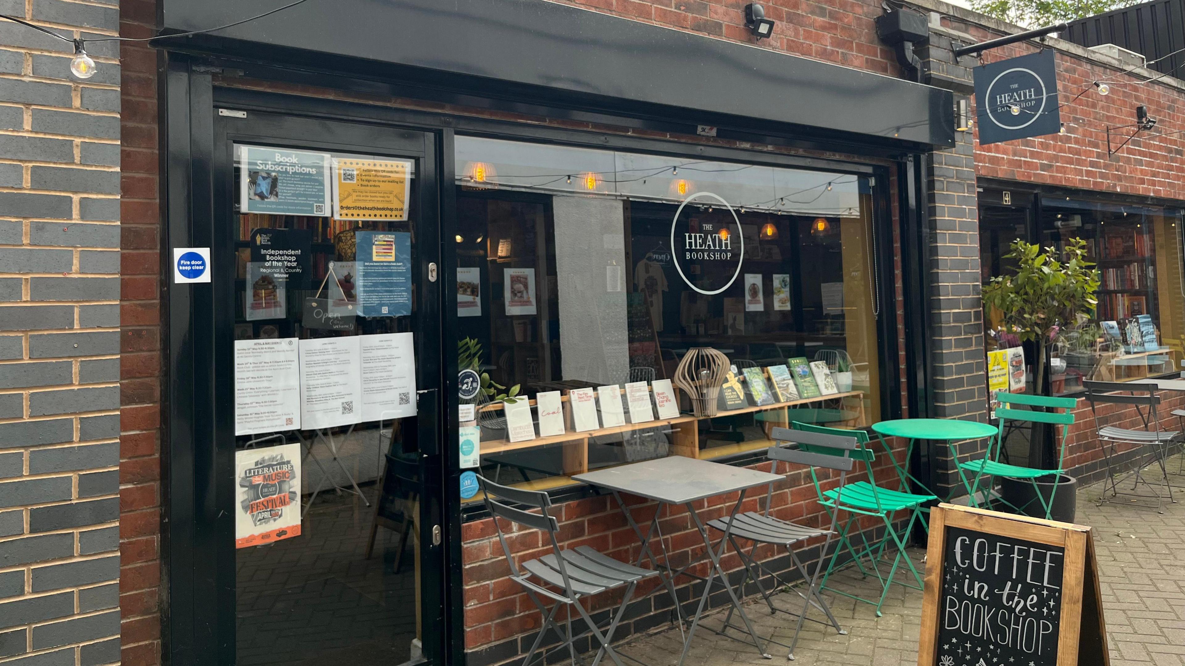 The outside of a shop with grey and green tables and chairs. There is a sign on the floor that says 'Coffee in the Bookshop'. Fairy lights are hanging in front of the shop door.