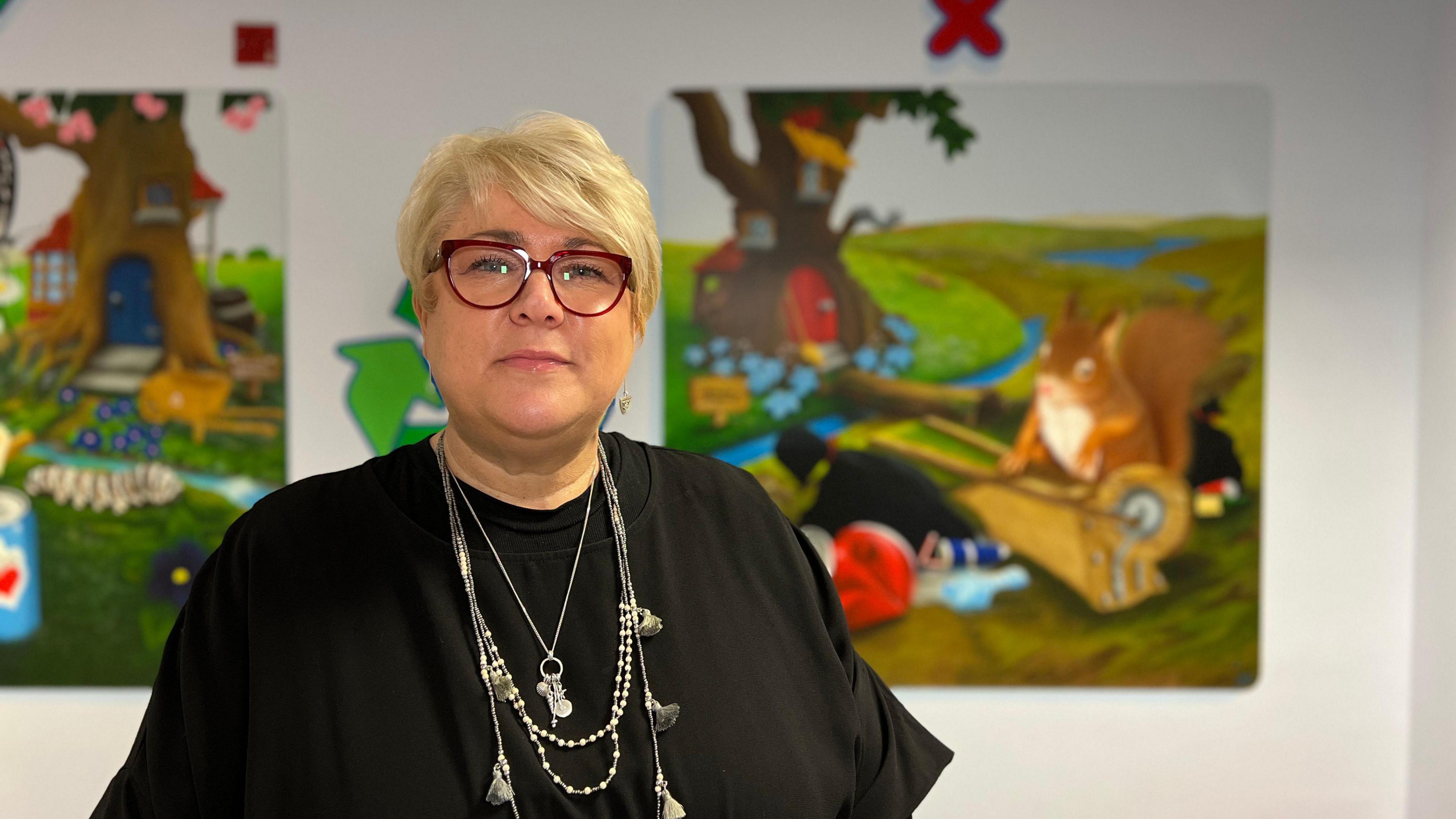 Michelle looks at the camera as she wears a black dress with silver necklaces on it. She has glasses with a thick, dark red frame and she has short blonde hair. Behind her are artworks of squirrels and rubbish.