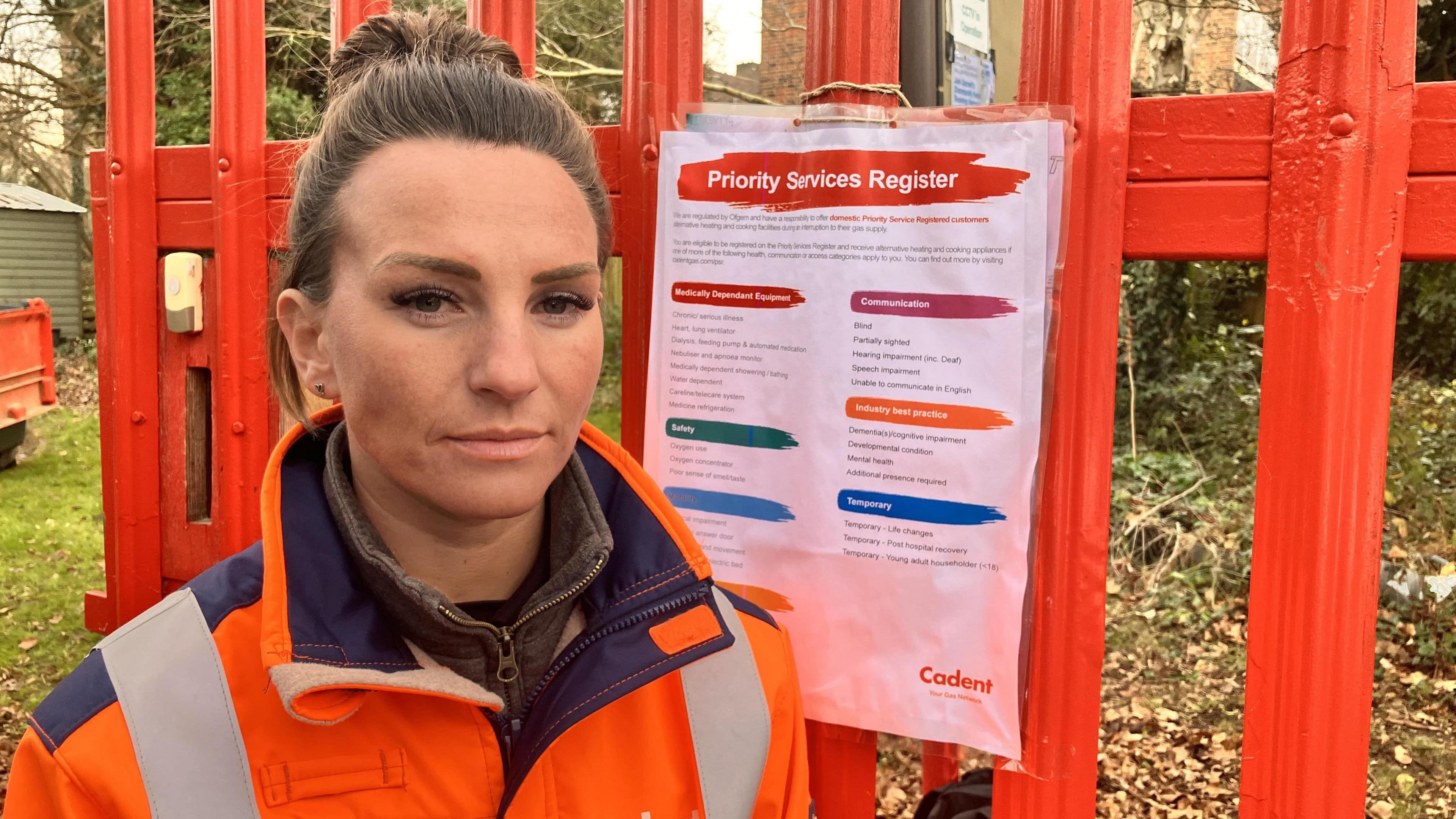 Vicky Grieve in an orange high-vis jacket standing in front of a red fence.