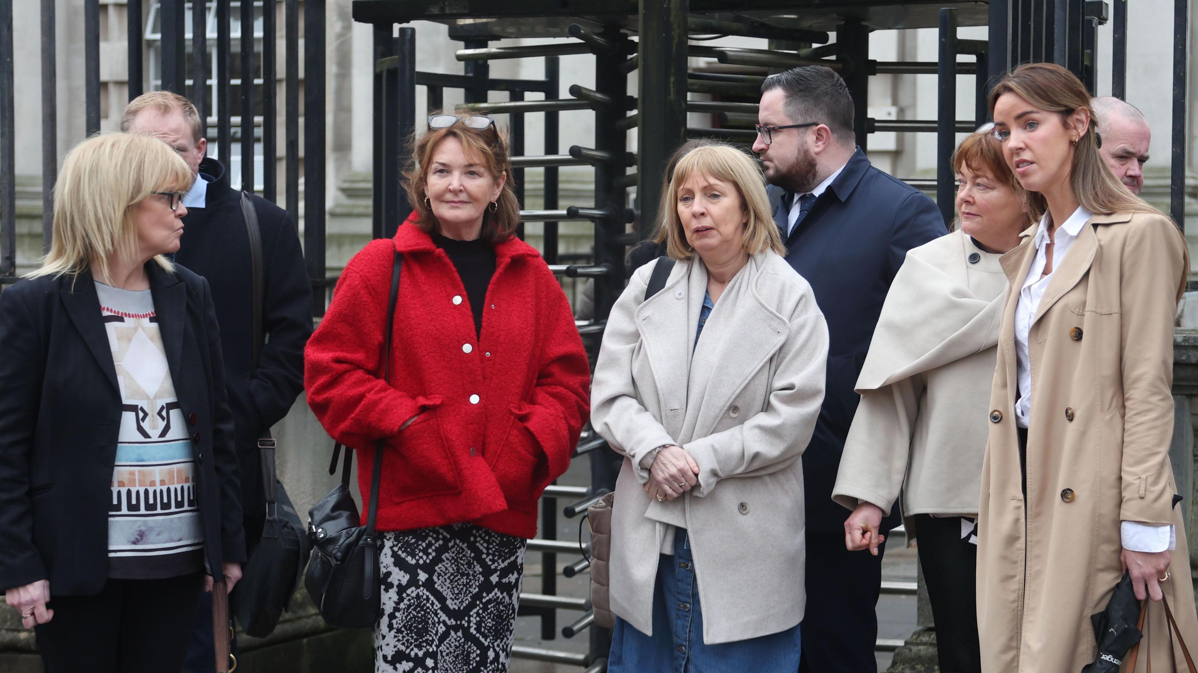 From left to right, a lady with medium length blonde hair wearing a patterned jumper and black blazer, a man is standing behind her but his face is not in focus. A lady stands next to him with medium length brown hair a red jacket, black top and snake print skirt, a woman with blonde hair stands next to her with a neutral colour coat. Behind this lady is a man with dark hair and facial hair wearing a coat and shirt and tie. Next to him is a lady with short hair and a white top, and next to her is a woman with long brown hair, white shirt and stone coloured trench coat. In the background is the black revolving gates into the courthouse.