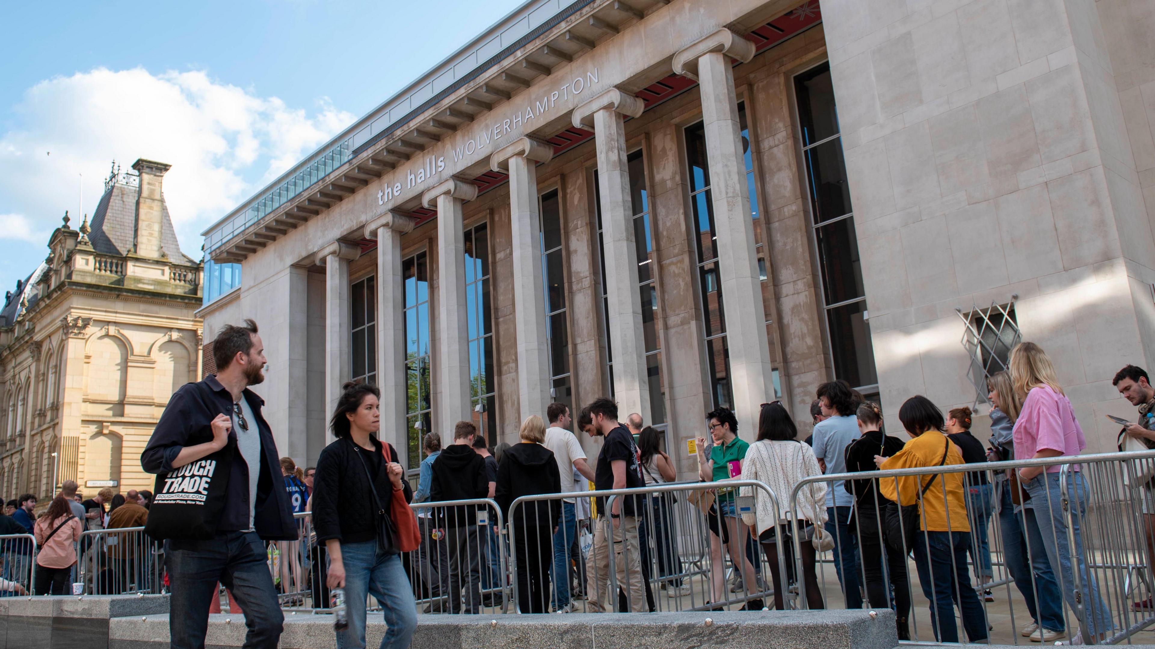 Vistors queuing alongside crowd barriers outside the entrance to the venue, some are looking at their phones. A man ans a woman look on as they pass in the opposite direction