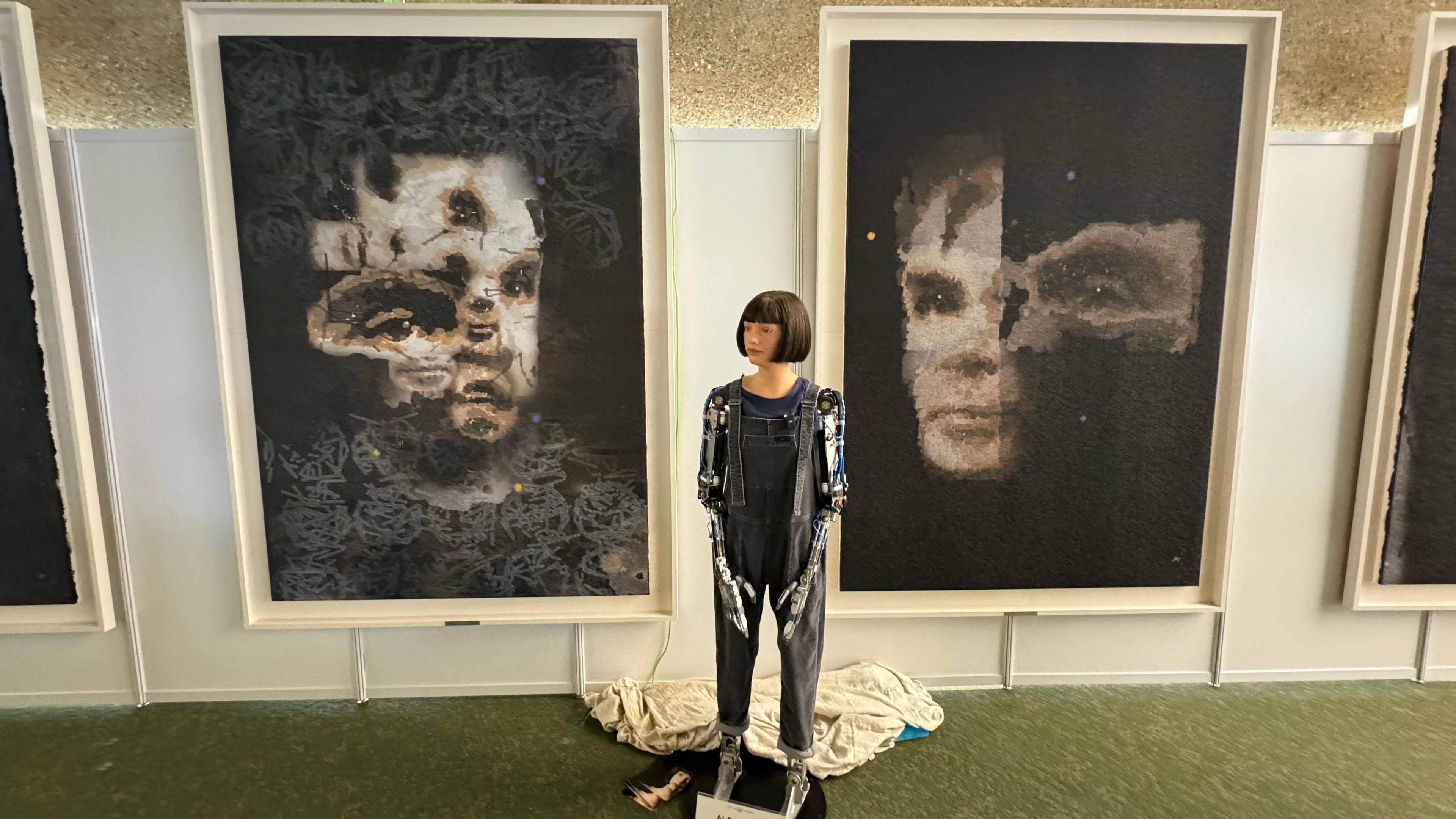 An AI robot, which looks like a woman with robotic arms stands in front of two paintings it has done. The paintings are of Alan Turing. 