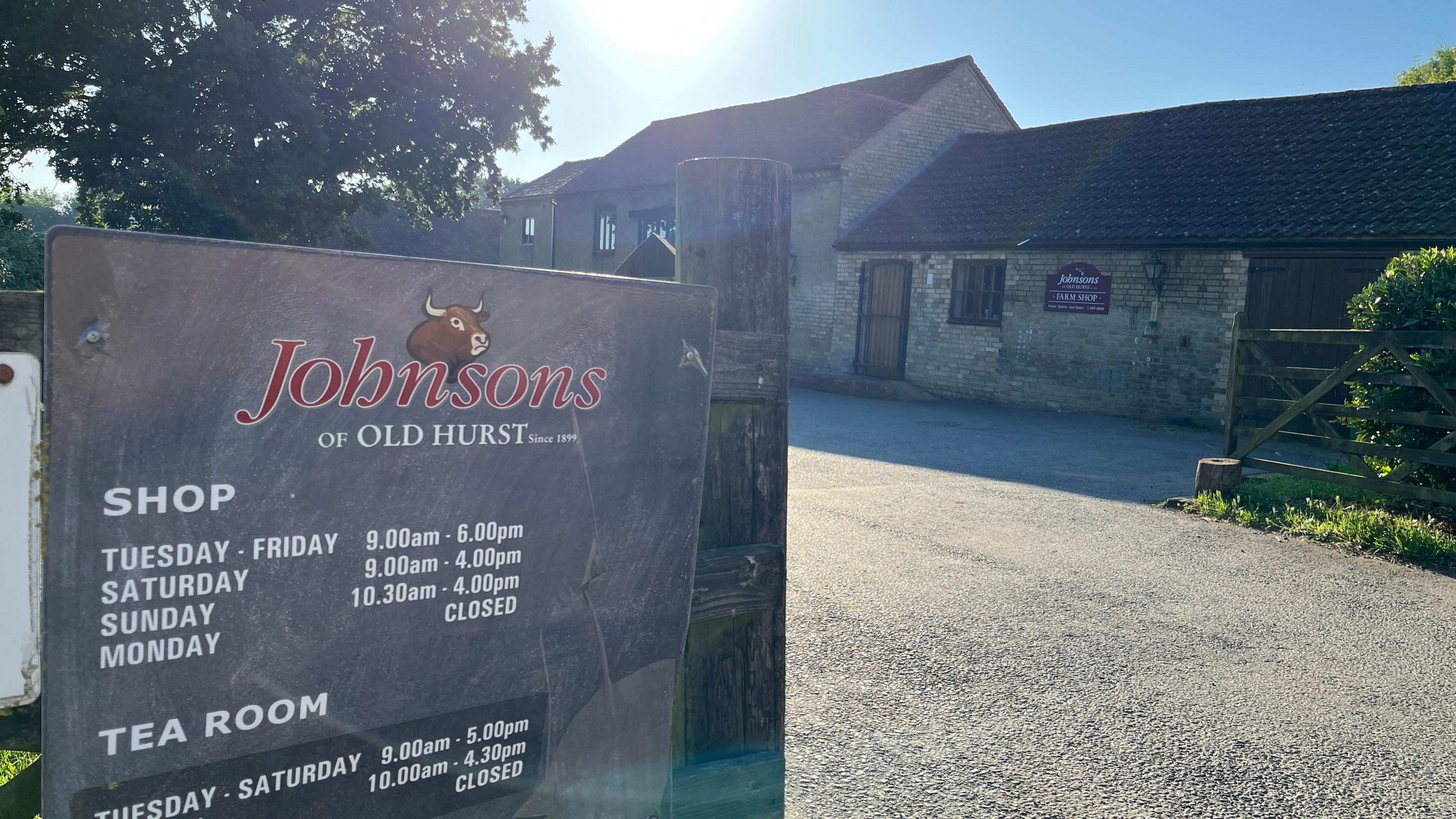 Johnsons of Old Hurst sign. Shows opening times. It is a bright sunny morning and there is a brick building in the background . 