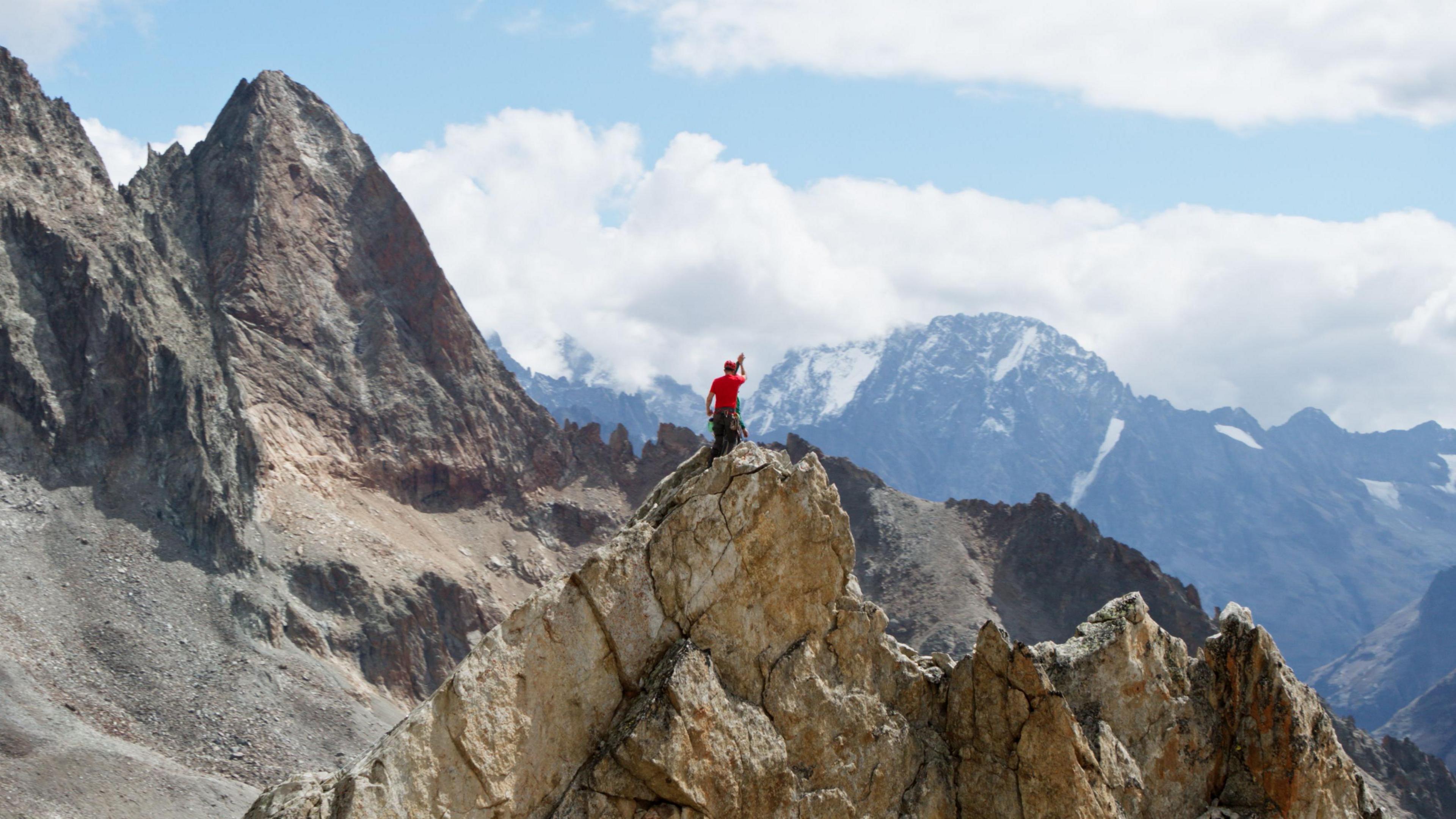 Ed Jackson standing on a mountain. The photograph is taken from afar - Ed is appearing as a small figure and can be seen in a red t-shirt. Mountains can be seen in the distance. 